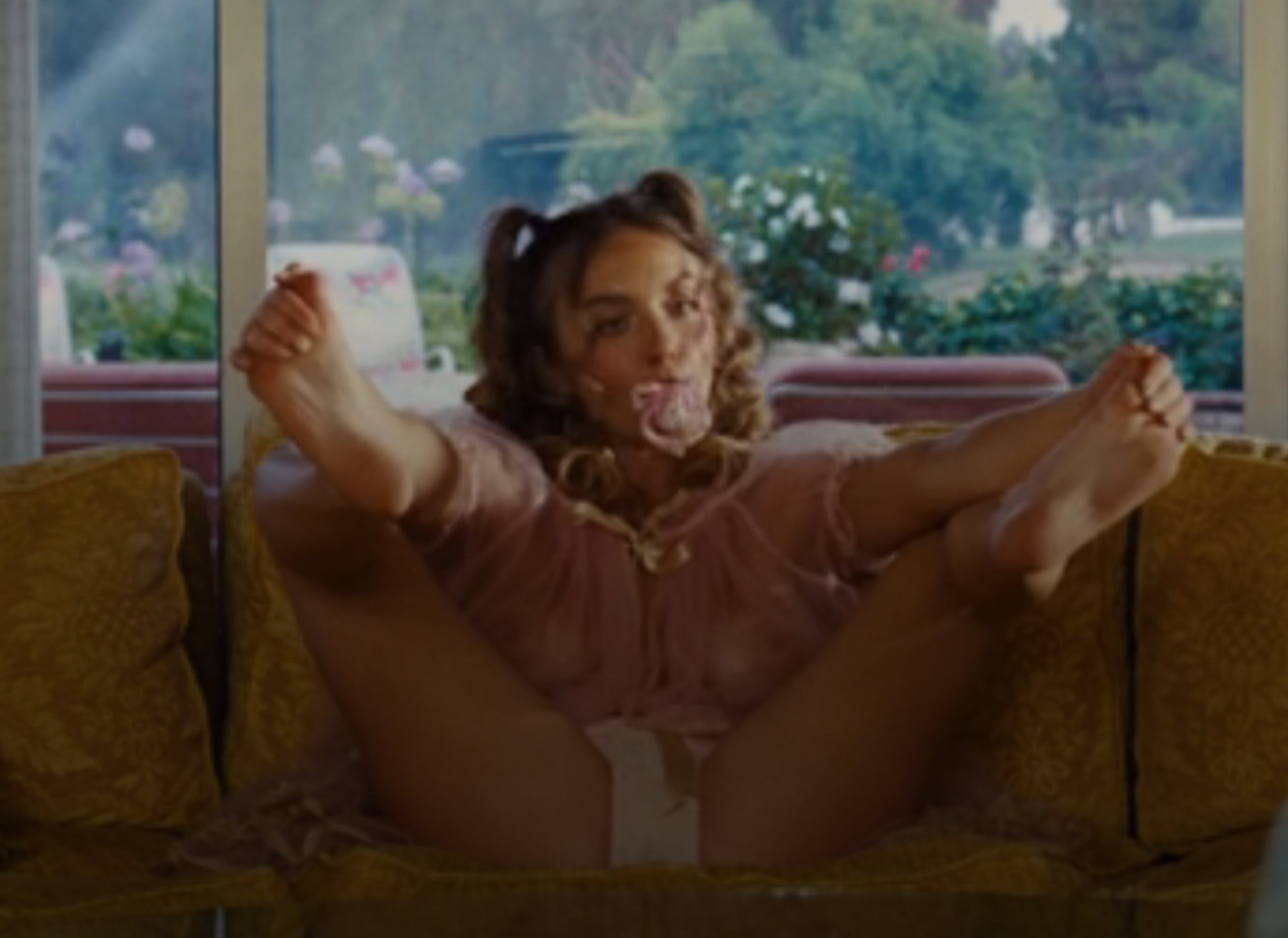 A idiosyncratic   is lounging playfully connected  a couch, blowing a bubblegum bubble, with trees disposable   done  ample  windows successful  the background