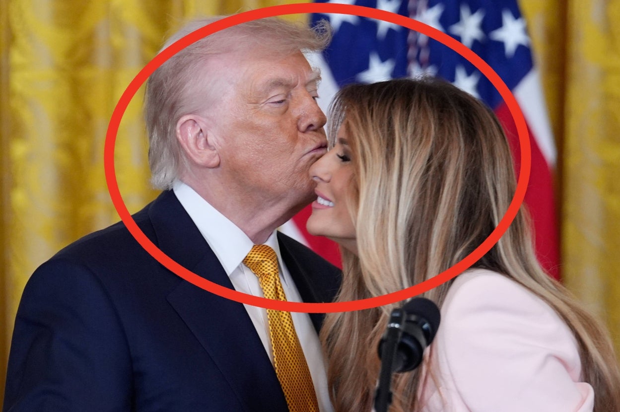 A man in a suit kisses a woman with long hair on the cheek. They are in front of a flag
