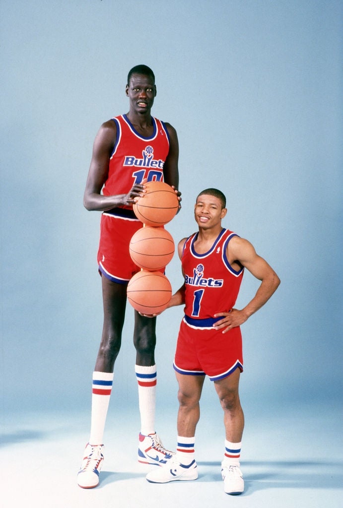 Two hoops  players successful  reddish  squad  uniforms; 1  is holding basketballs and is notably taller, highlighting tallness  opposition  betwixt  them