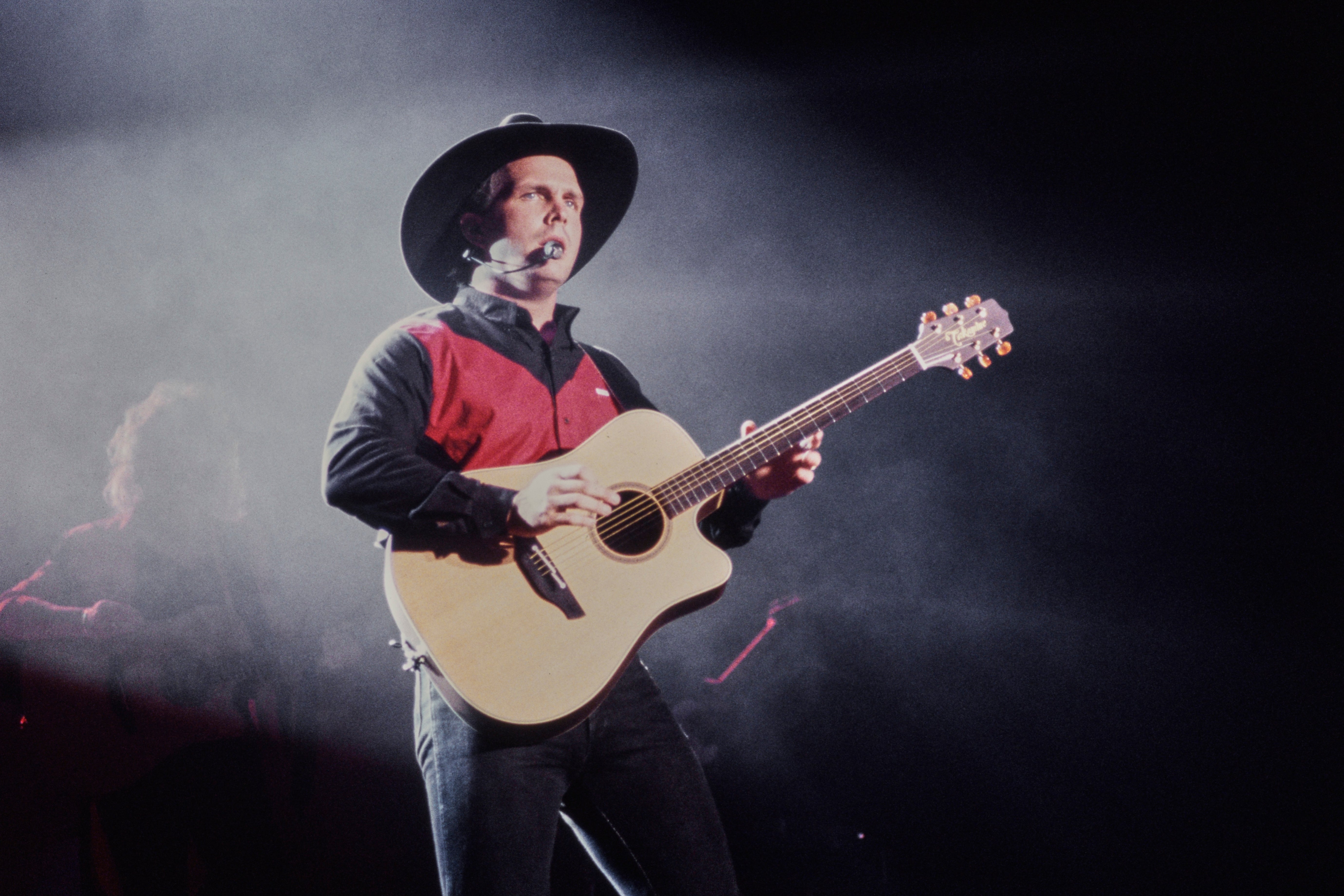 Singer successful cowboy chapeau and reddish garment performs energetically connected signifier with an acoustic guitar, acceptable against melodramatic lighting