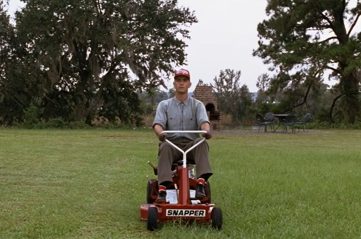 Forrest Gump mowing a large lawn on a ride-on lawn mower, surrounded by trees and outdoor furniture in the background