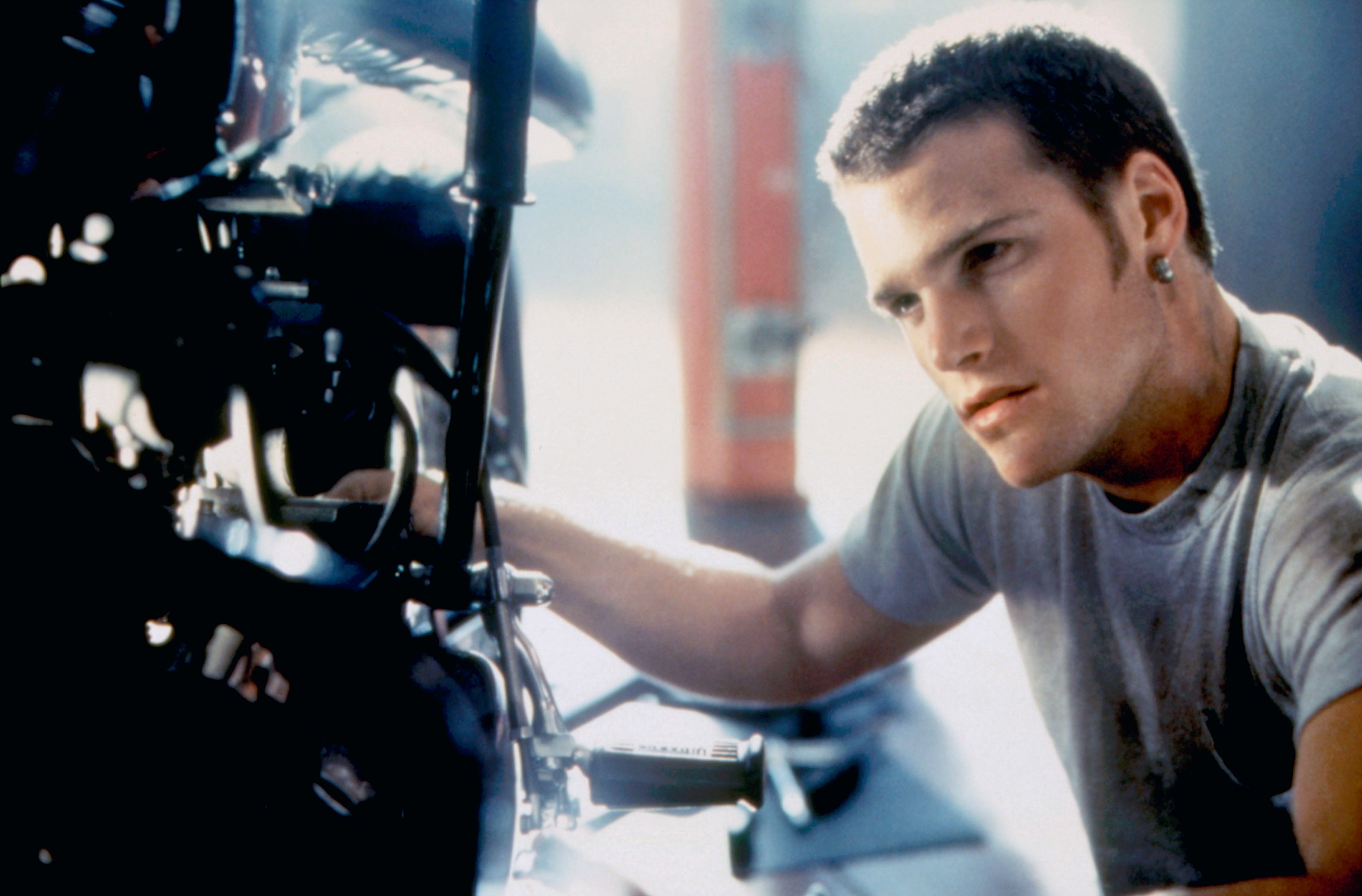 Person examining a motorcycle engine, wearing a t-shirt and an earring, looking focused and intent connected the task