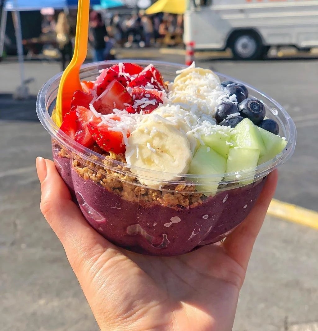 Smoothie bowl topped with sliced strawberries, bananas, blueberries, and melon in a hand. Granola and coconut flakes add texture