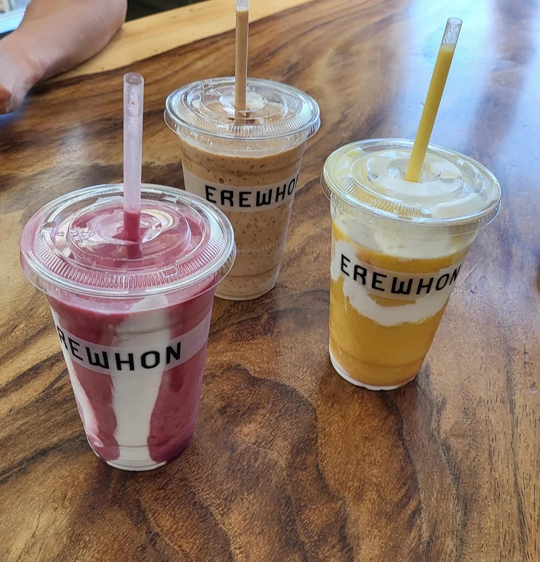 Three smoothies on a wooden table, each in a cup labeled "Erewhon." They are strawberry, mocha, and mango flavors from left to right