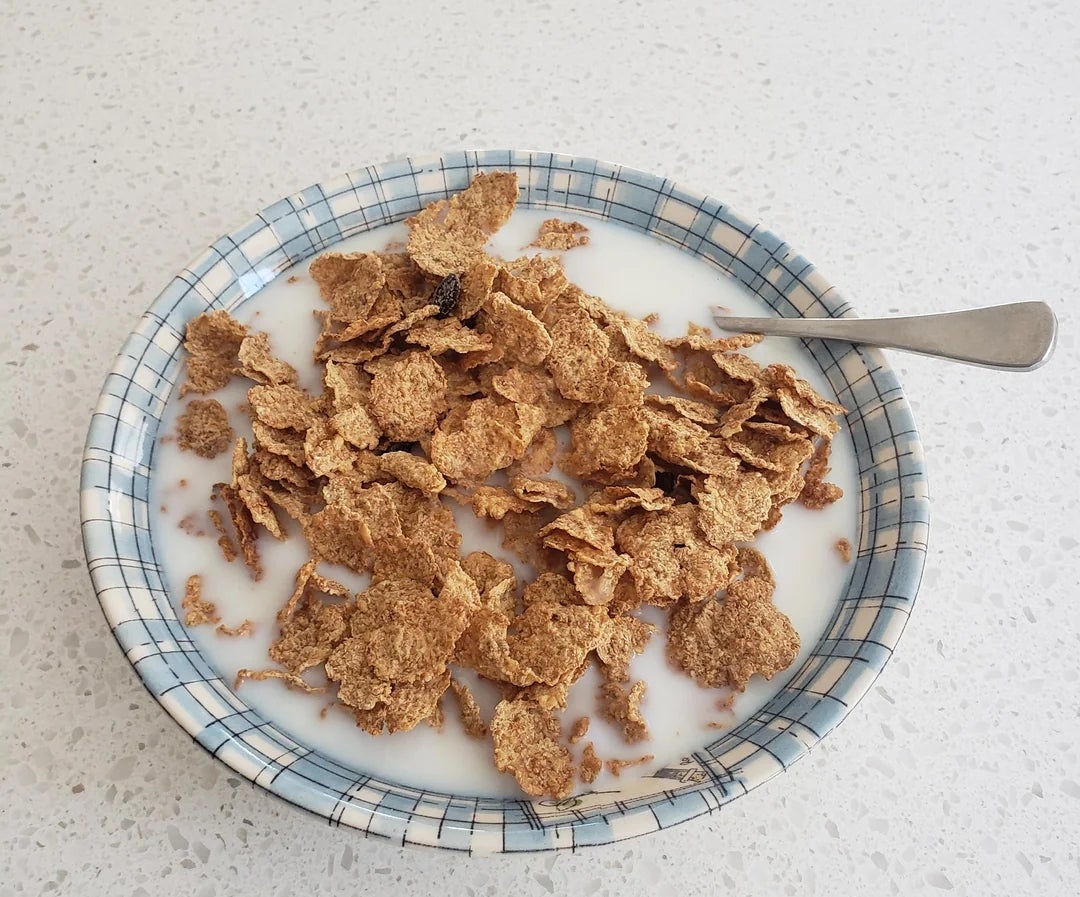 A bowl of bran flakes with milk on a checkered plate, with a spoon resting inside