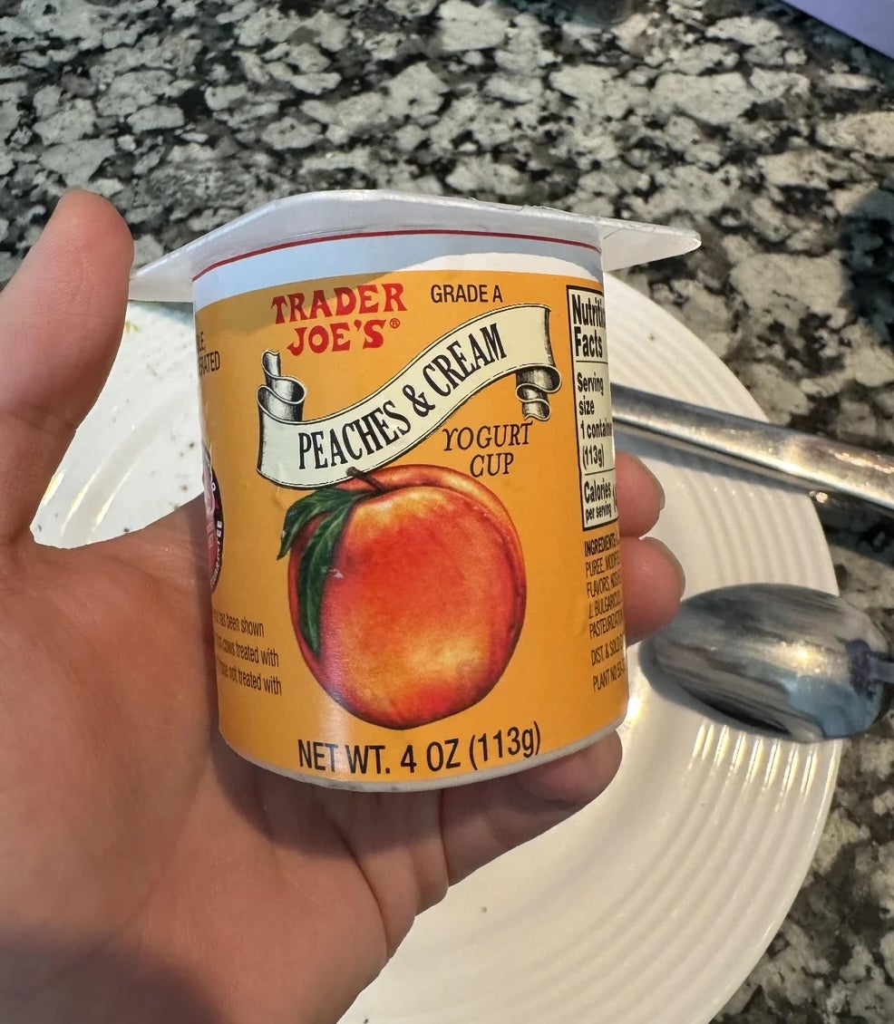 Person holding a Trader Joe's Peaches &amp;amp; Cream yogurt cup over a kitchen counter with a spoon nearby