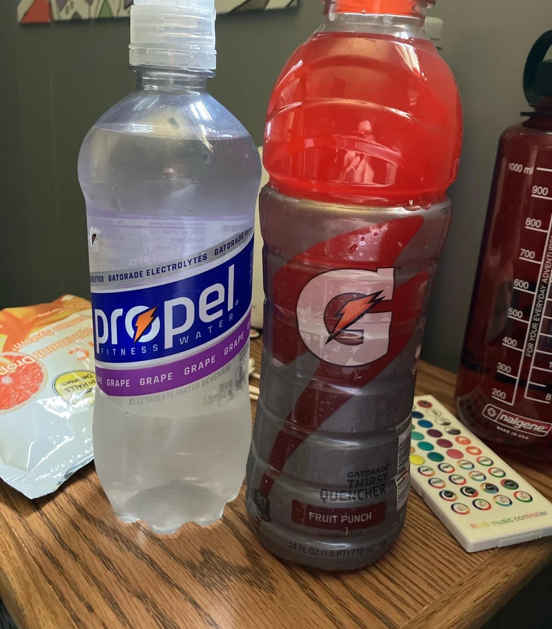 Two sports drinks on a table: a Propel fitness water bottle and a Gatorade Fruit Punch bottle. In the background, a large water bottle and snacks