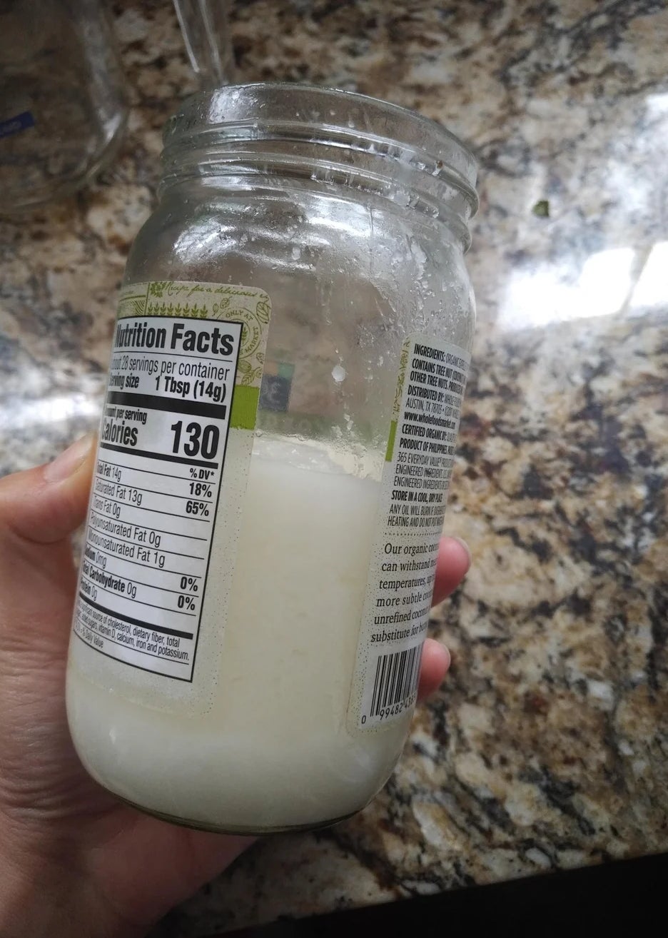 Hand holding a jar of coconut butter with visible nutrition facts label. Jar is placed on a marble countertop