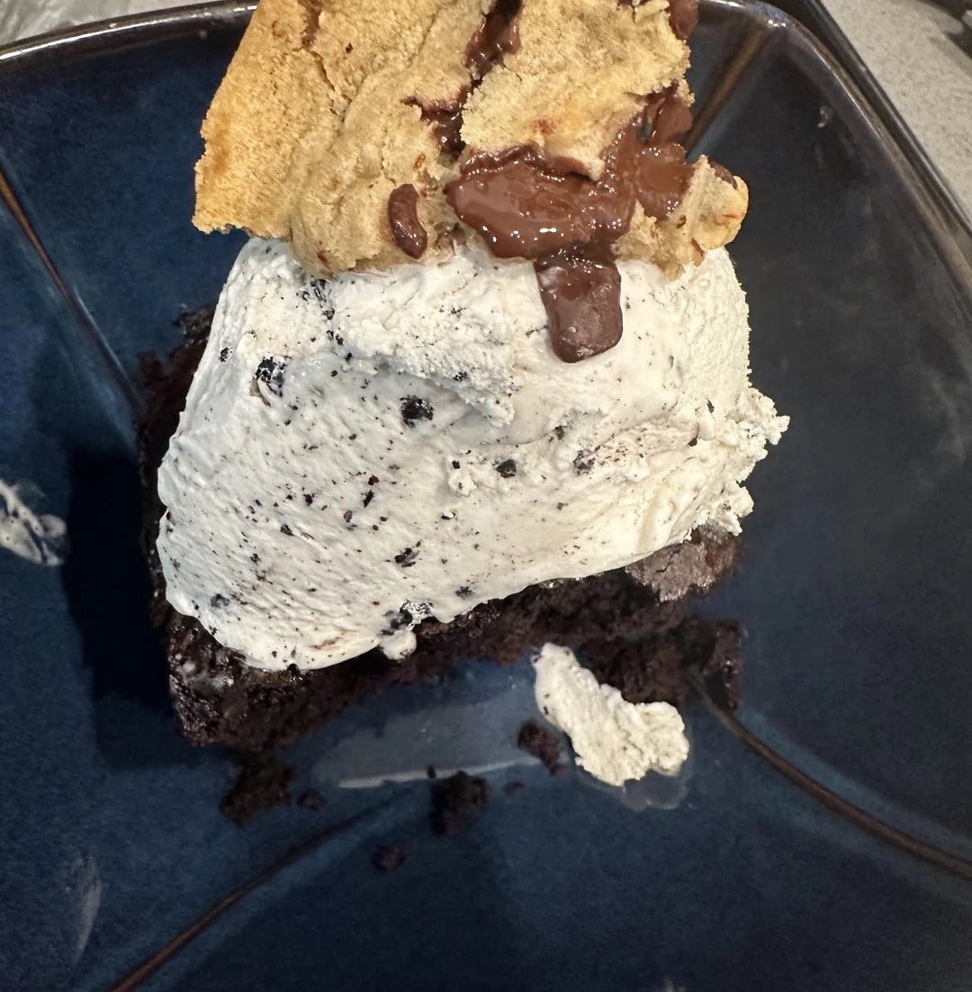A decadent dessert with a scoop of cookies and cream ice cream on chocolate cake, topped with a chocolate chip cookie
