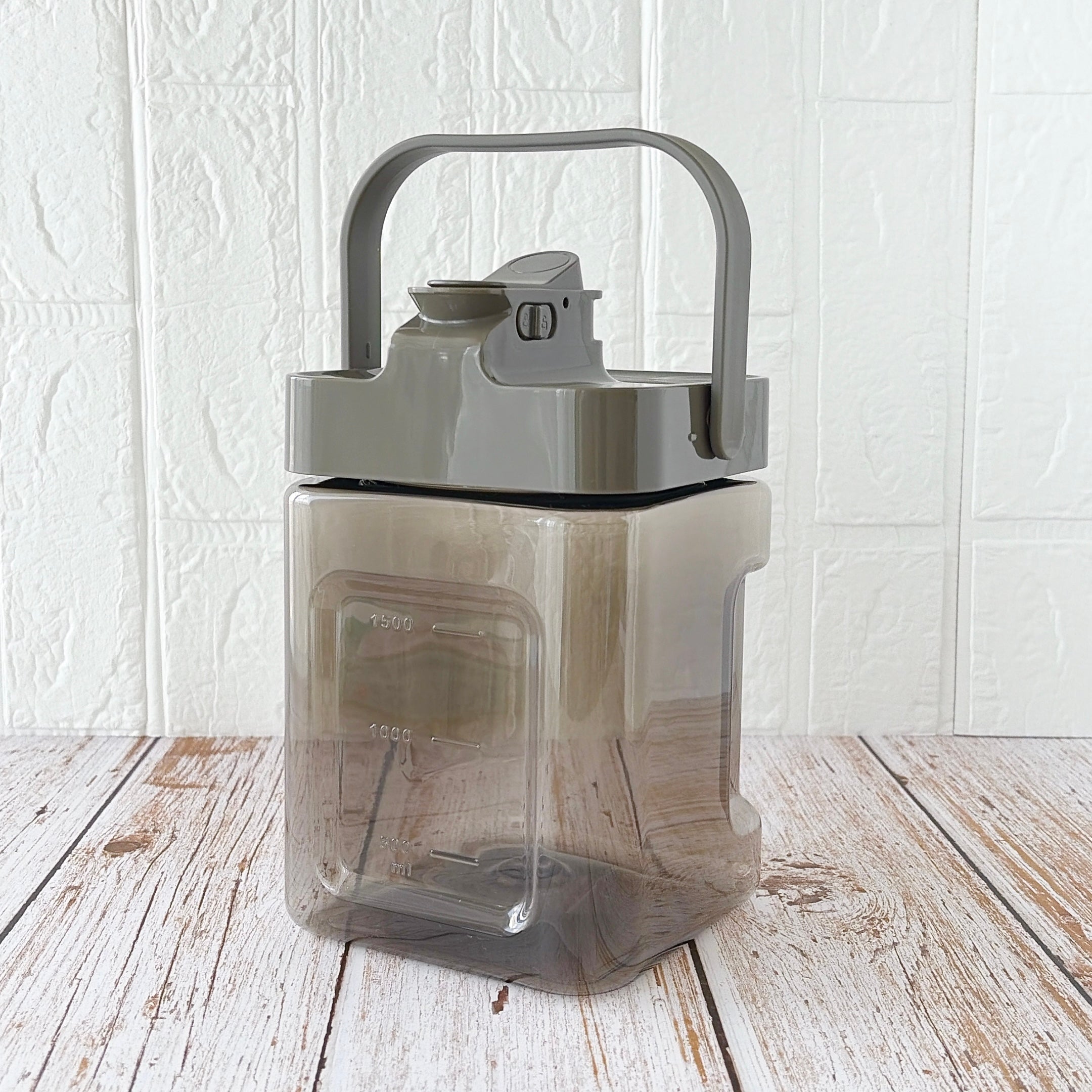 3COINS（スリーコインズ）「ドリンクサーバー：2.3L／KITINTO」透明な密閉容器が、木目調の床に置かれている。持ち手と蓋が付いているデザイン。