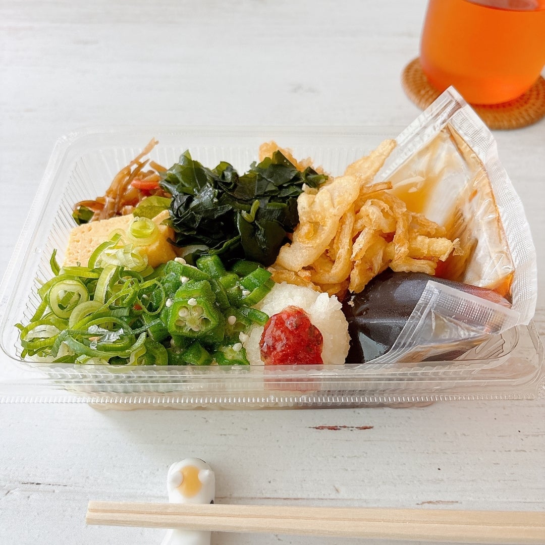 丸亀製麺の「梅おろしオクラうどん弁当」の全体的な様子。天ぷら、野菜、わかめなどが入った食材が透明なプラスチック容器に盛り付けられている。