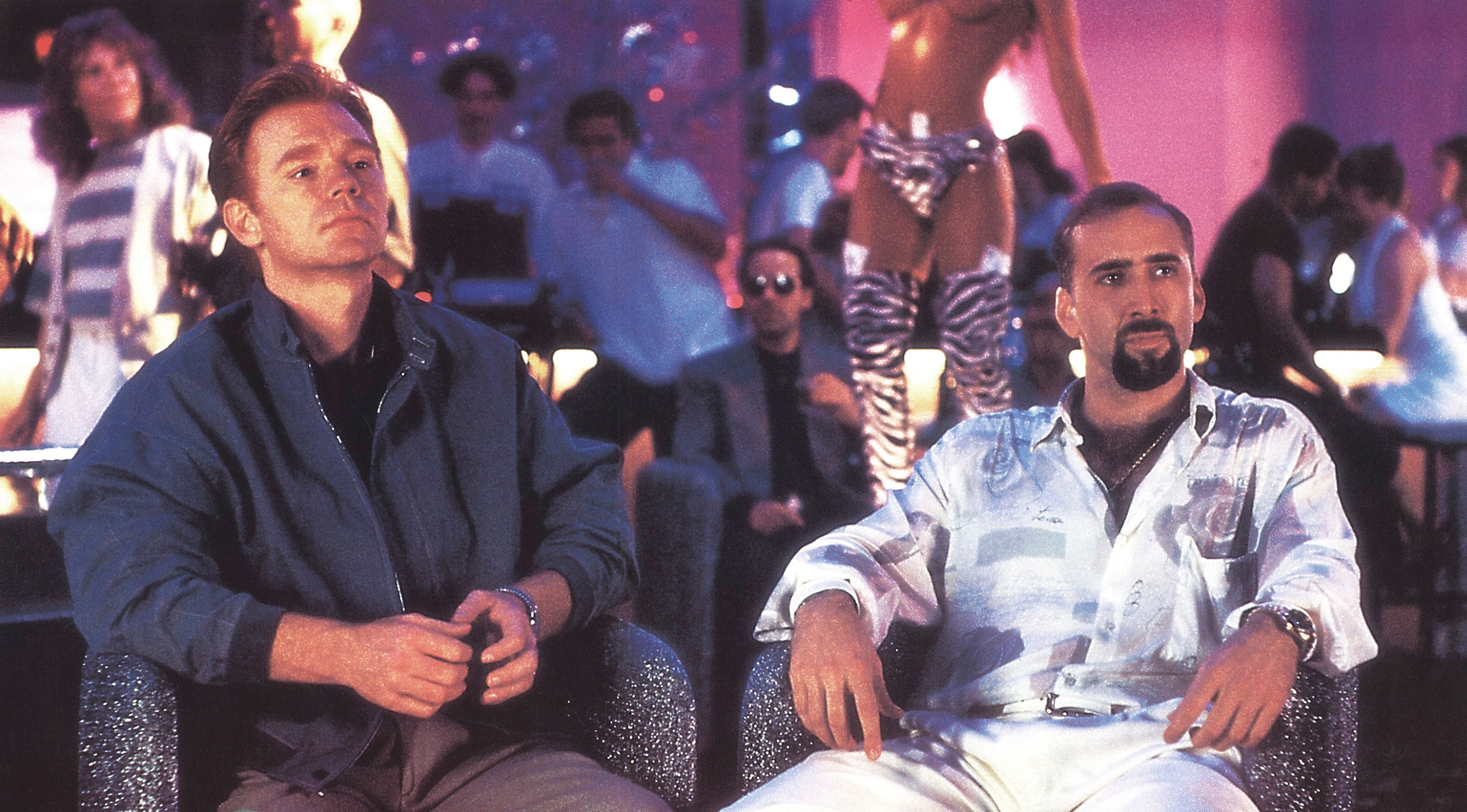 Two men seated successful a lively nine setting. One wears a jacket, the different a patterned, casual shirt. Background features clubgoers and a dancer