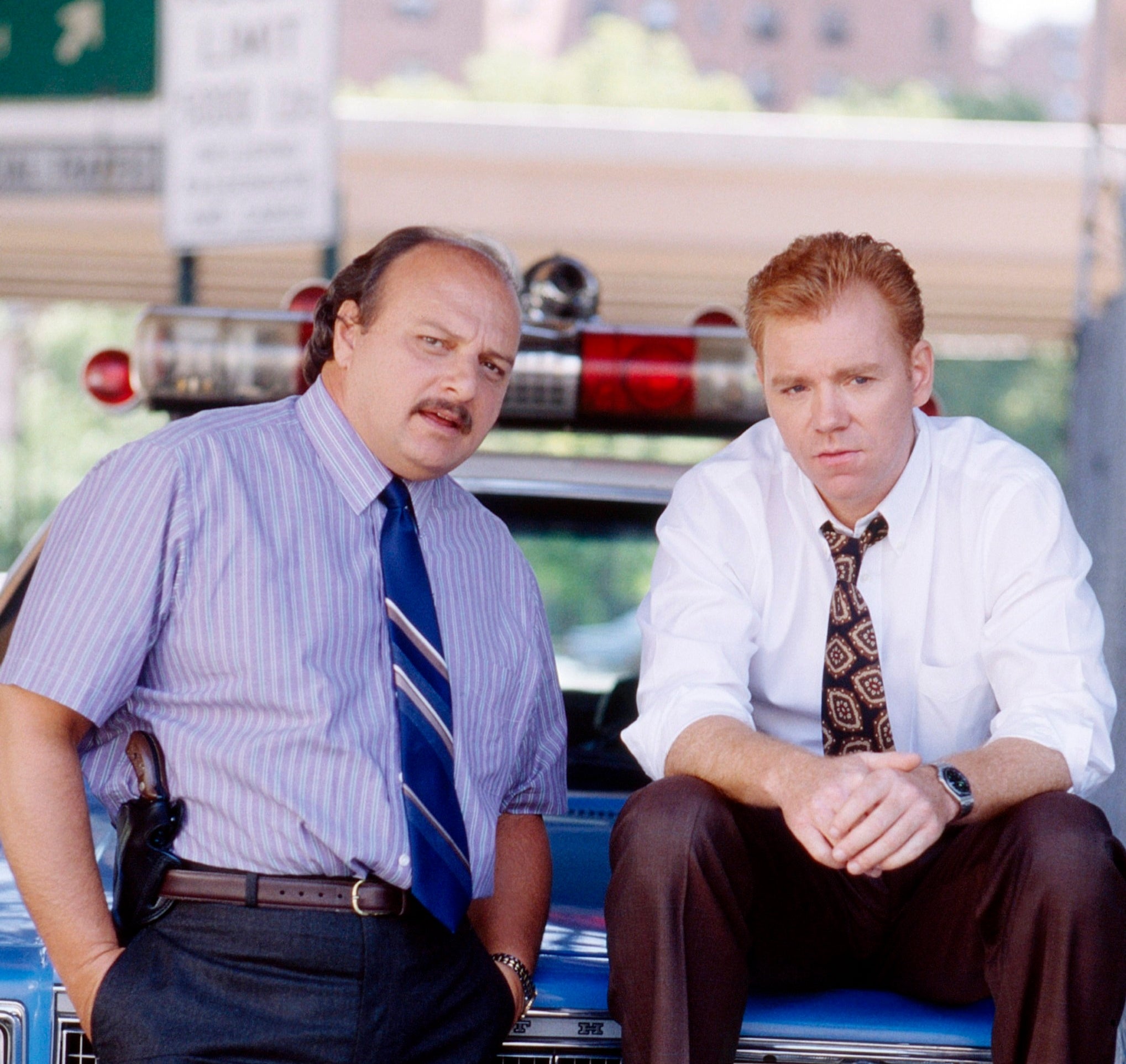 Two actors portraying detectives thin connected a car, dressed successful 1990s concern attire. One wears a necktie and shirt; the different sports a mustache and patterned tie