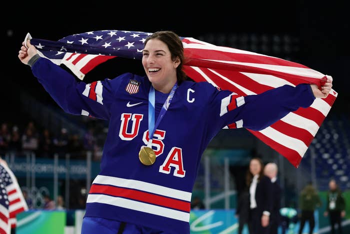 Athlete wearing a USA hockey jersey and gold medal celebrates with an American flag draped over her shoulders on an ice rink