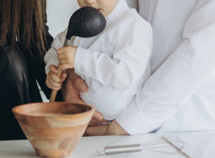 子供が大きなスプーンを持ち、大人に抱えられながらキッチンで木のボウルに向かっている。