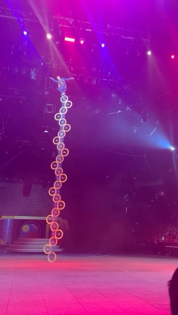 Performer balances connected a tall, vertical stack of chairs nether signifier lights, showcasing an acrobatic stunt successful a ample venue