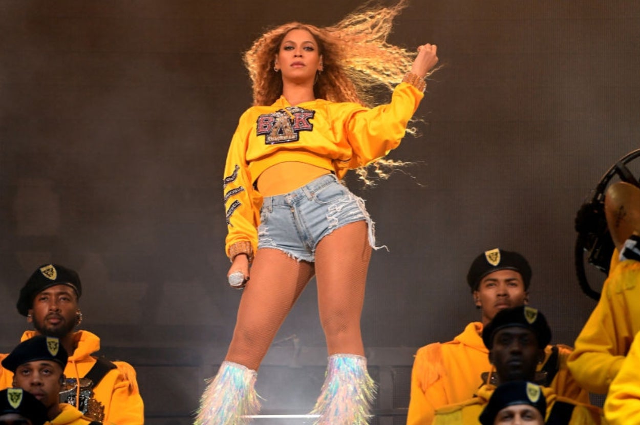 Person on stage in a cropped sweatshirt, ripped denim shorts, and knee-high boots with fringe, performing energetically