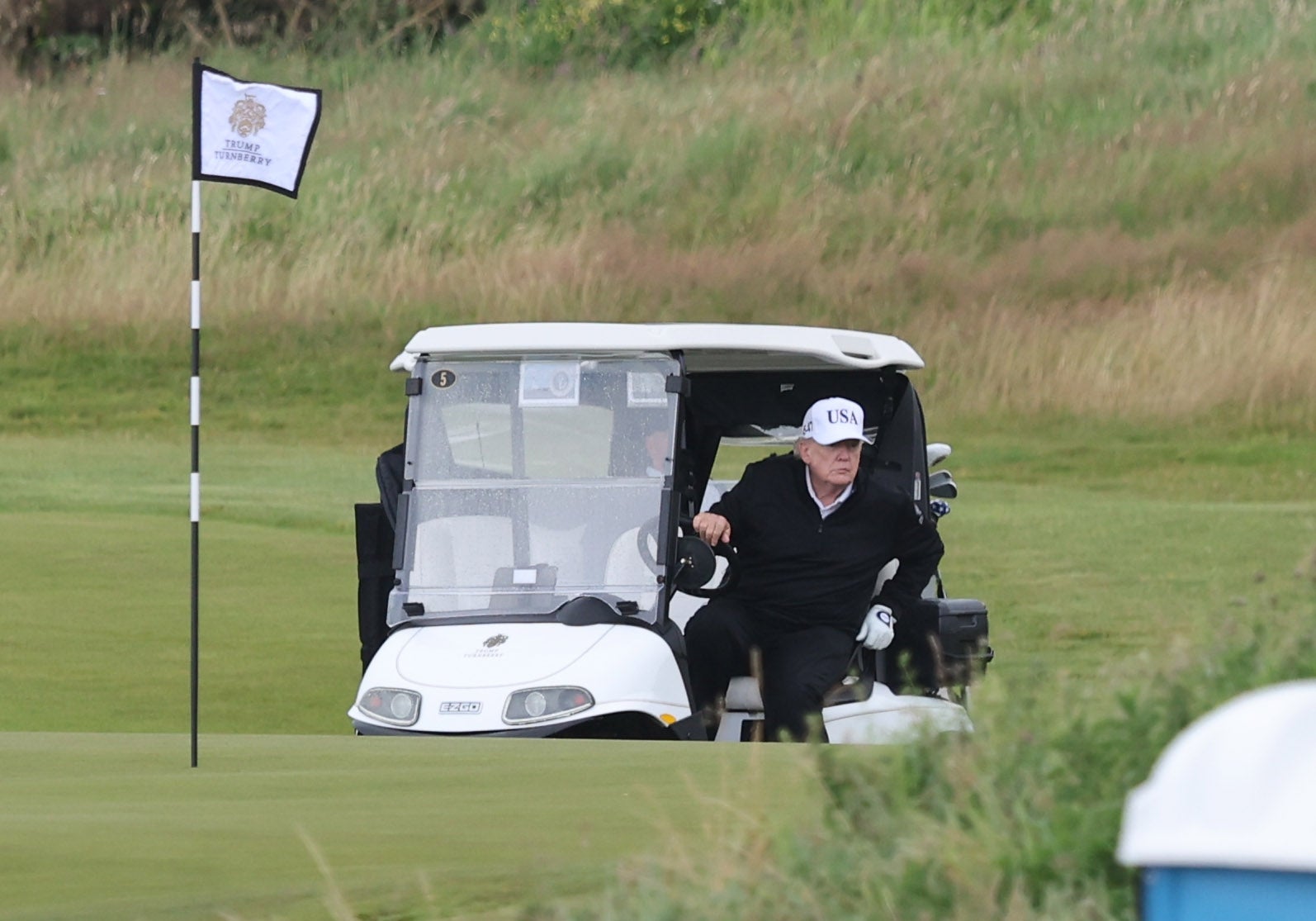 En person i caps og jakke som sitter i en golfbil på en golfbane, nær et flagg