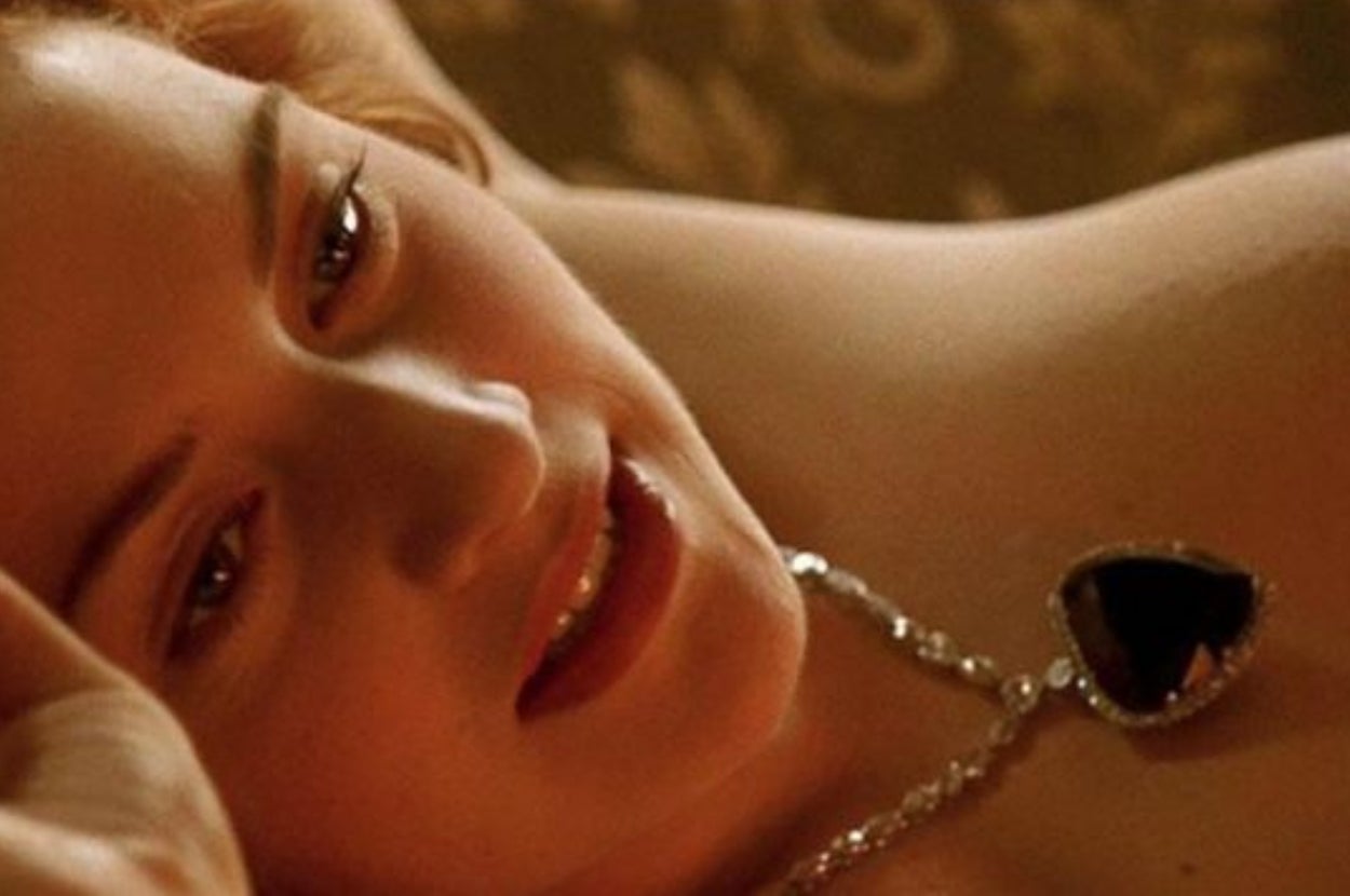 A person reclines, wearing a necklace with a large heart-shaped pendant, looking upwards thoughtfully