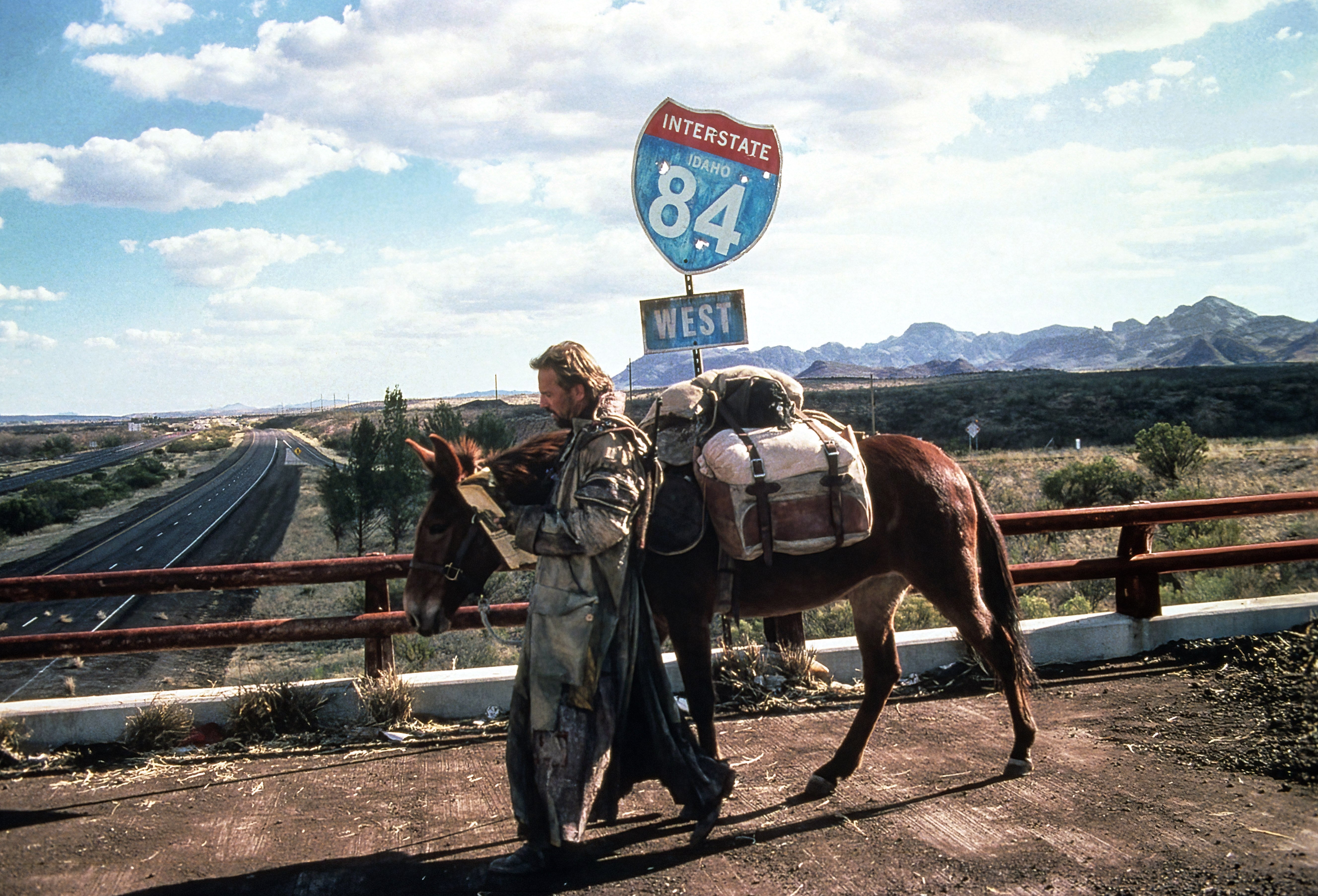 Person successful rugged covering walking a mule connected a bridge, nether an Interstate 84 West sign, with a road and mountains successful the background