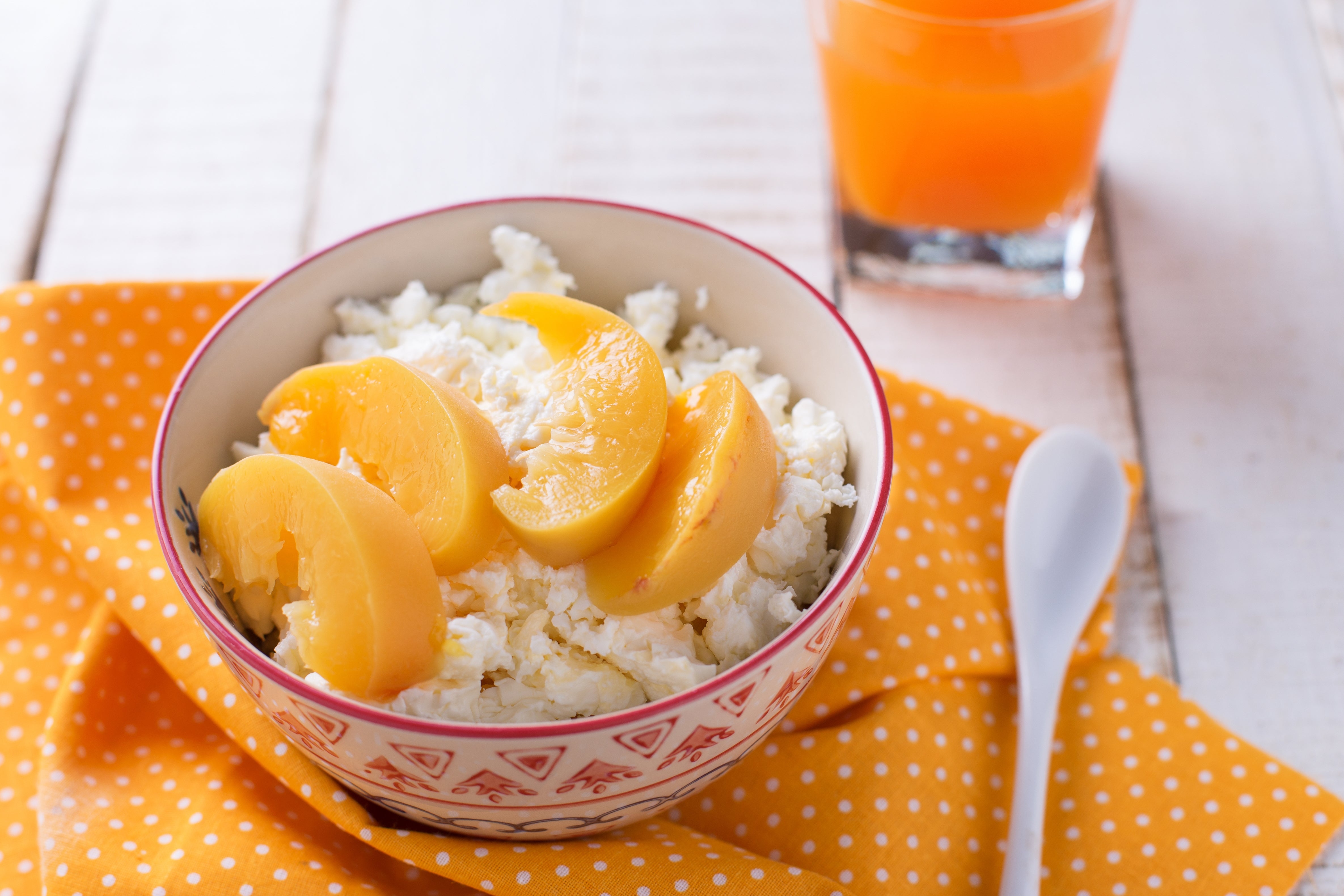 Bowl of cottage cheese topped with peach slices on a polka dot cloth, with a spoon and a glass of orange juice in the background