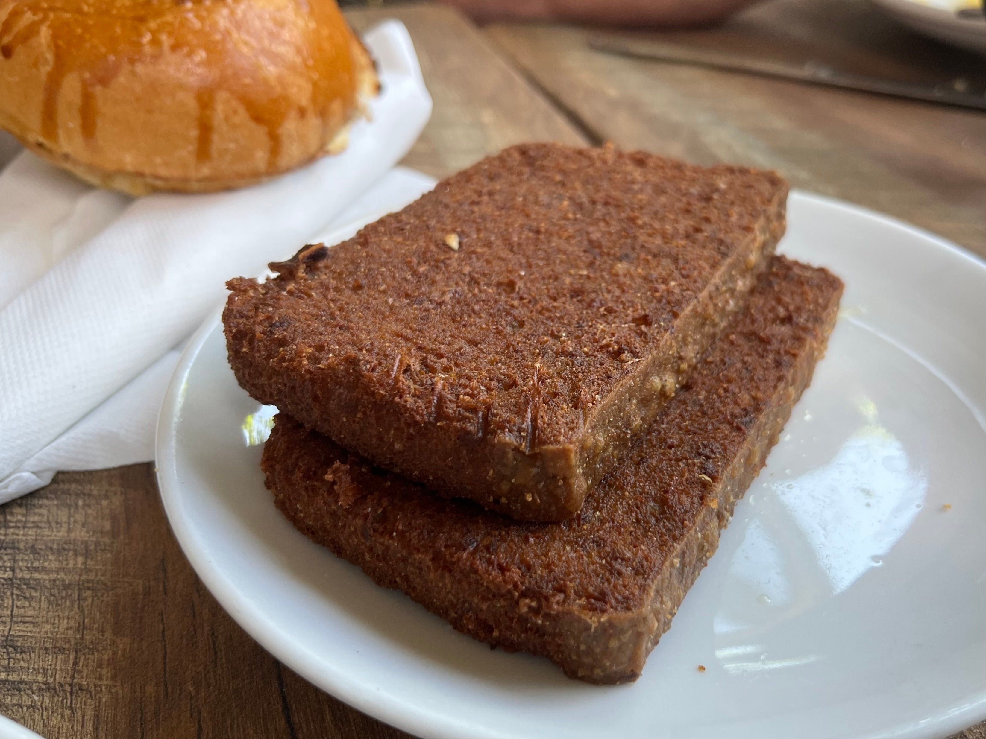 Two slices of scrapple on a white plate, next to a bun on a napkin