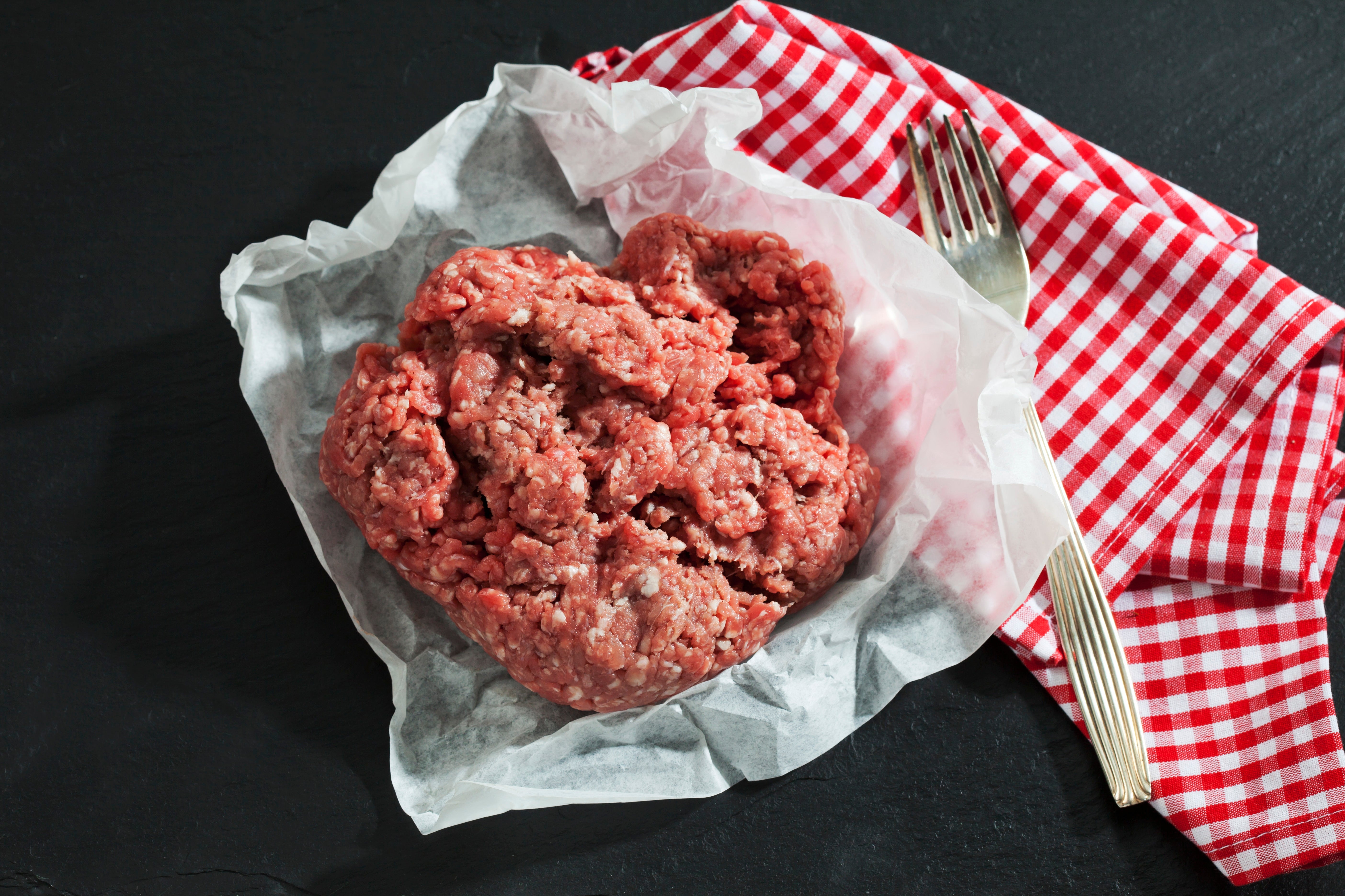 Ground beef on white paper next to a red-and-white checkered cloth and a fork on a dark surface