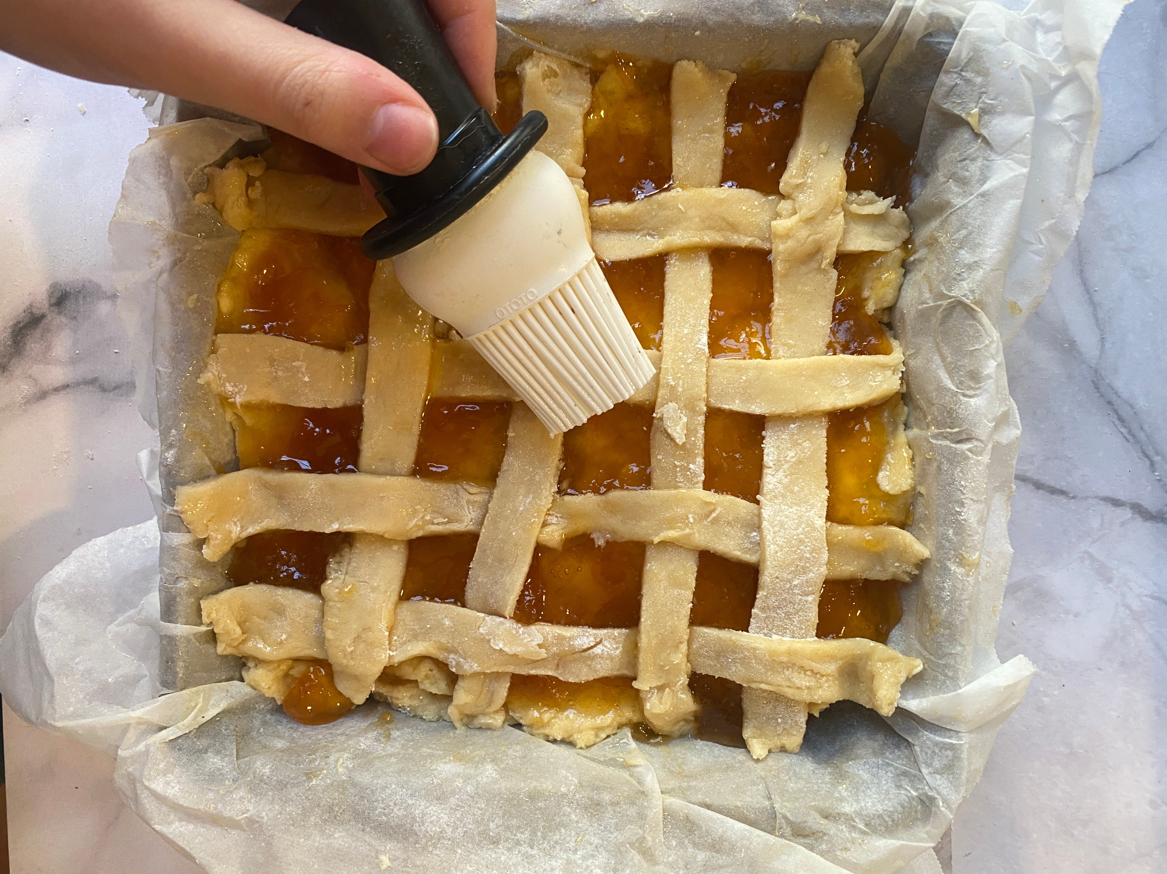 Hand brushes lattice pastry top of a homemade pie in a parchment-lined square pan