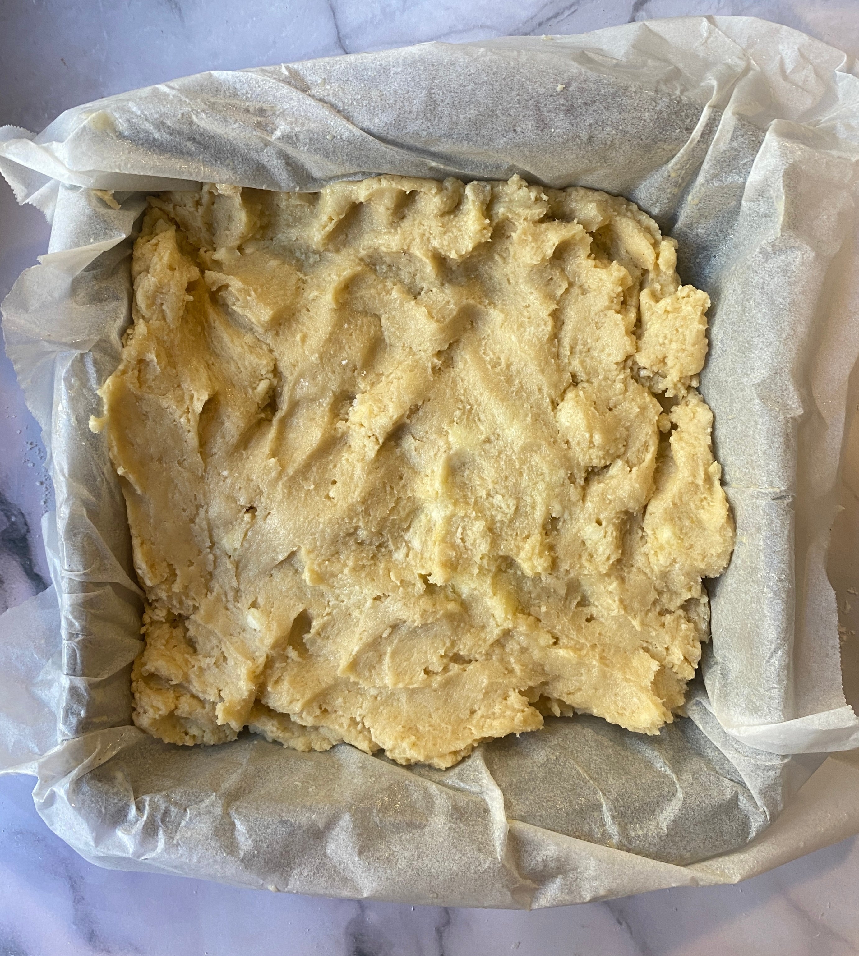Unbaked square cake batter spread in a parchment-lined baking pan on a marble surface