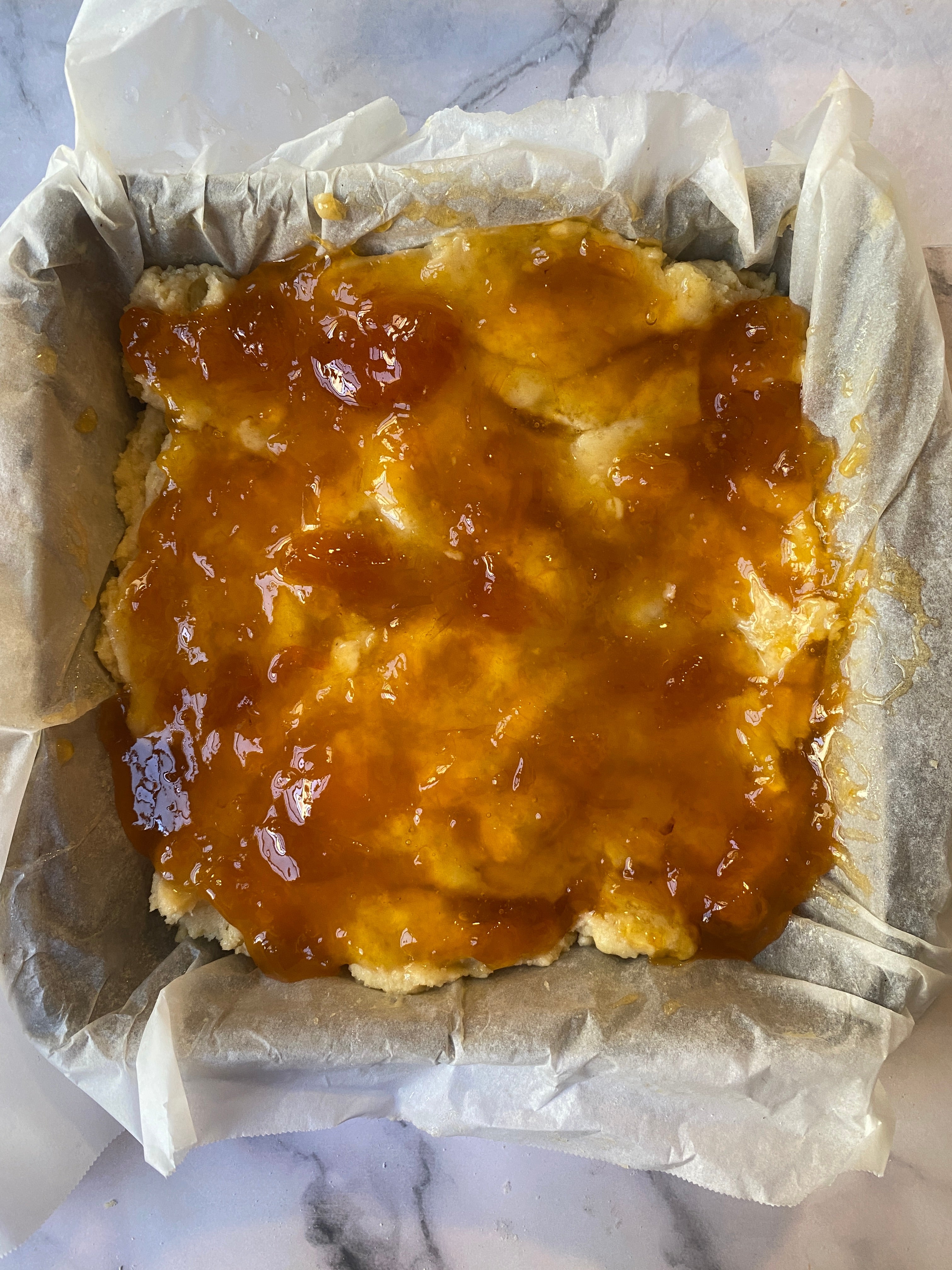 Baked dessert with a crumbly base, topped with a glossy fruit jam, in a square pan lined with parchment paper
