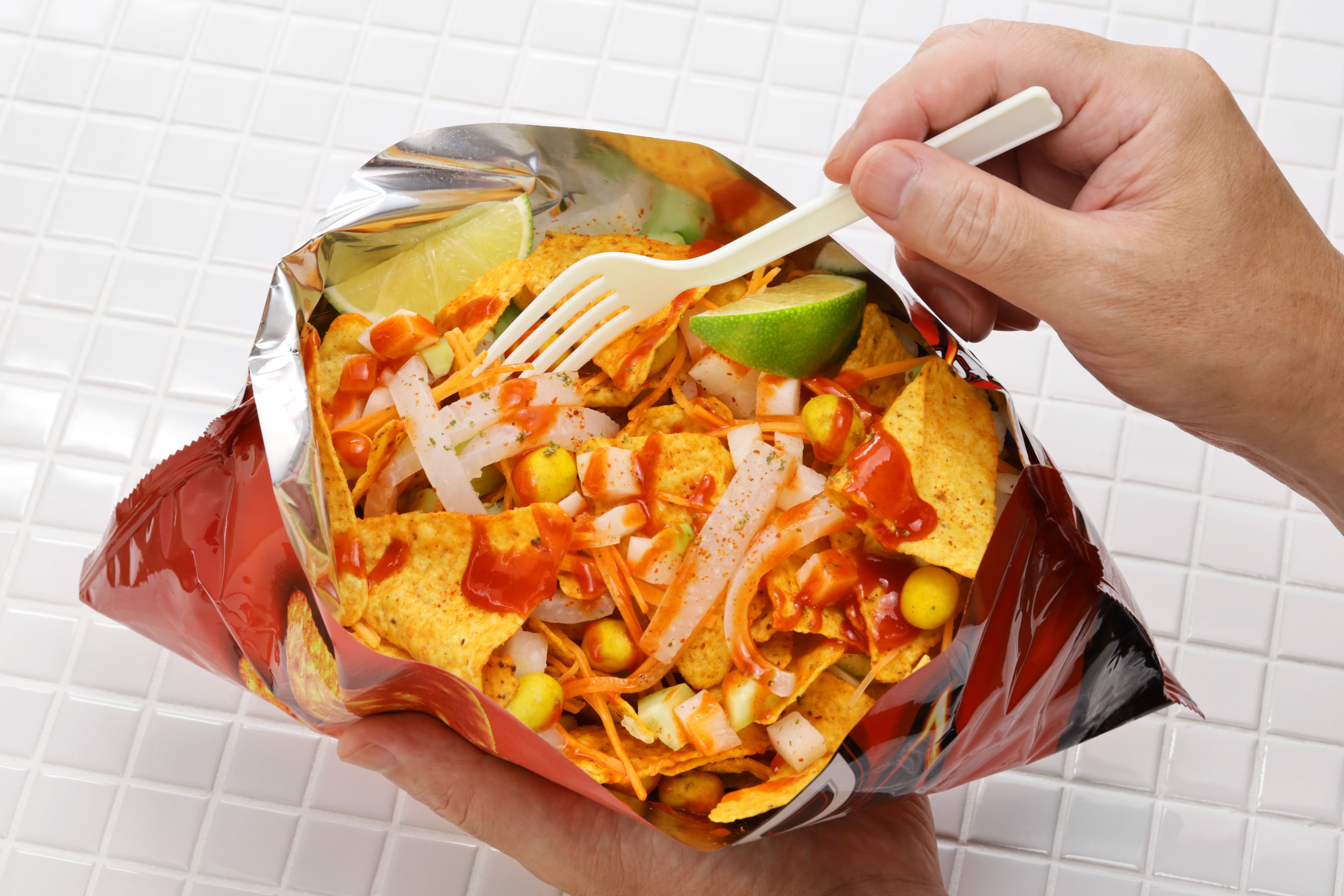 A person's hand holding a fork above a bag of tostilocos with corn, onions, lime slice, and hot sauce on a tiled surface