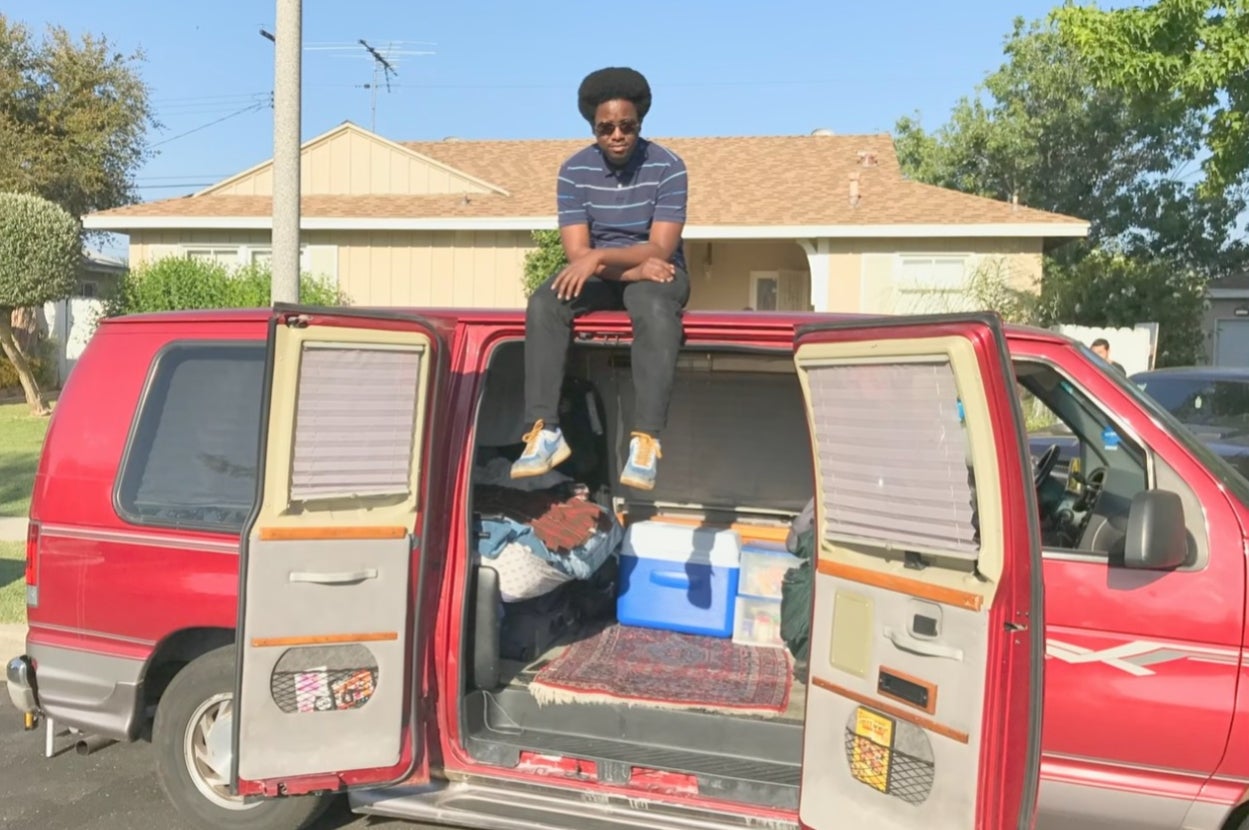 Person sitting on top of a van filled with belongings, parked in a residential neighborhood