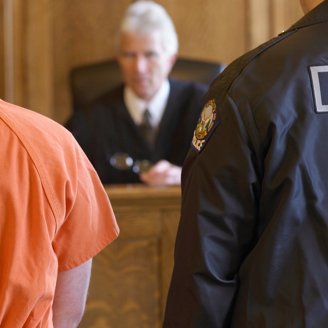Person in an orange jumpsuit facing a judge in a courtroom. Police officer stands nearby