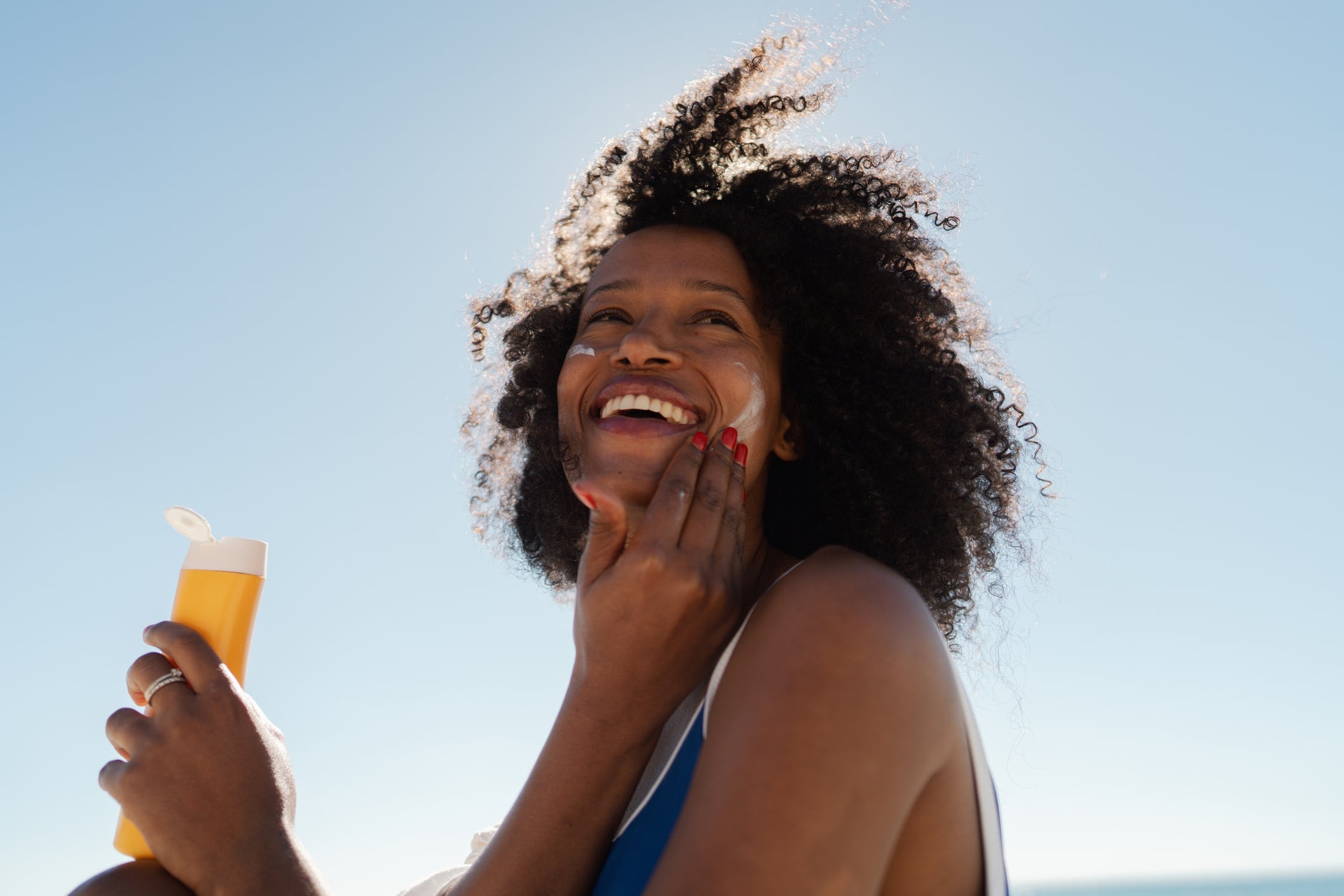 Person smiling while applying sunscreen at the beach, holding sunscreen bottle, wind blowing hair