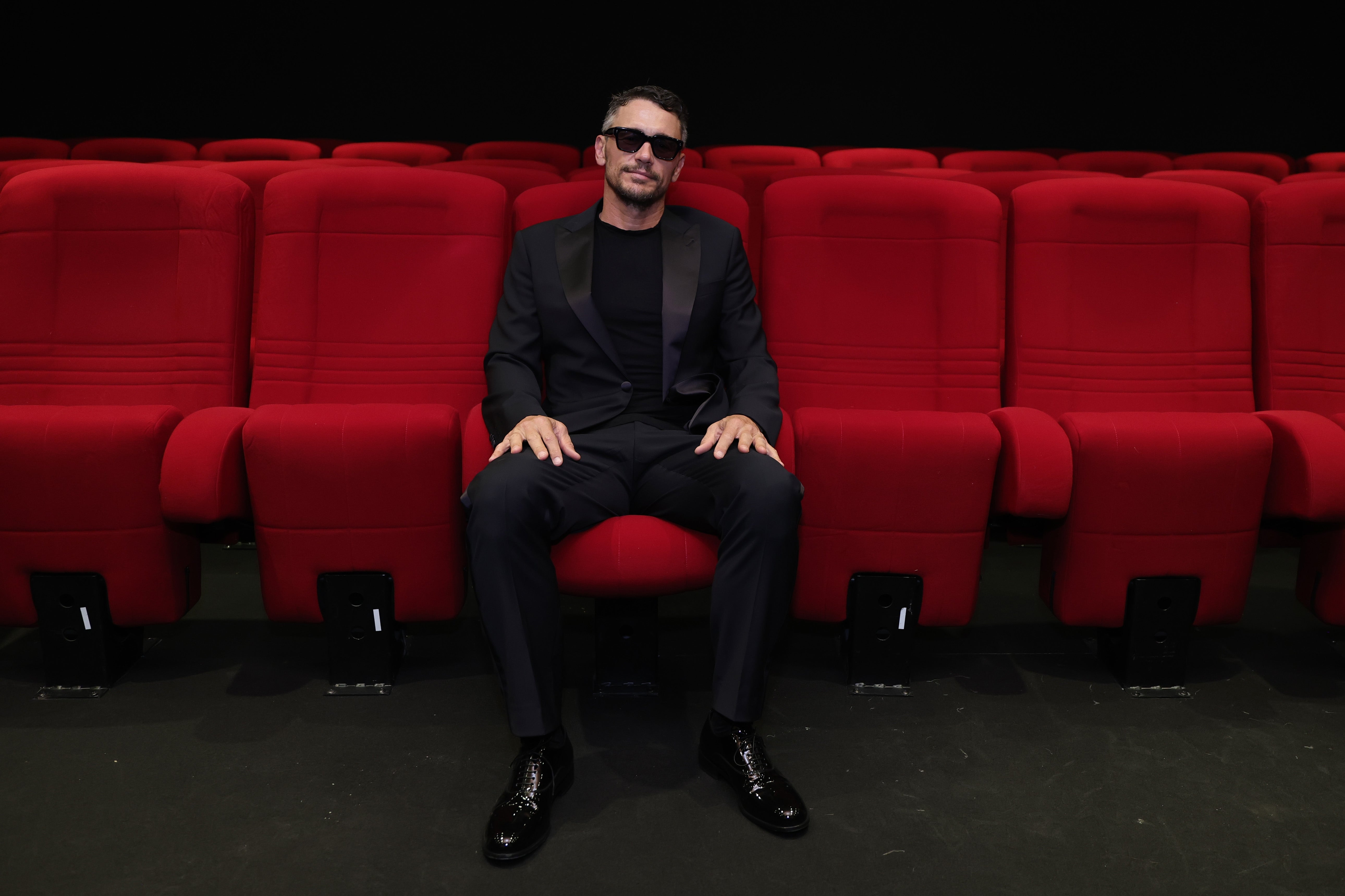 Mann i mørk dress med solbriller sitter alene i et teater med røde seter