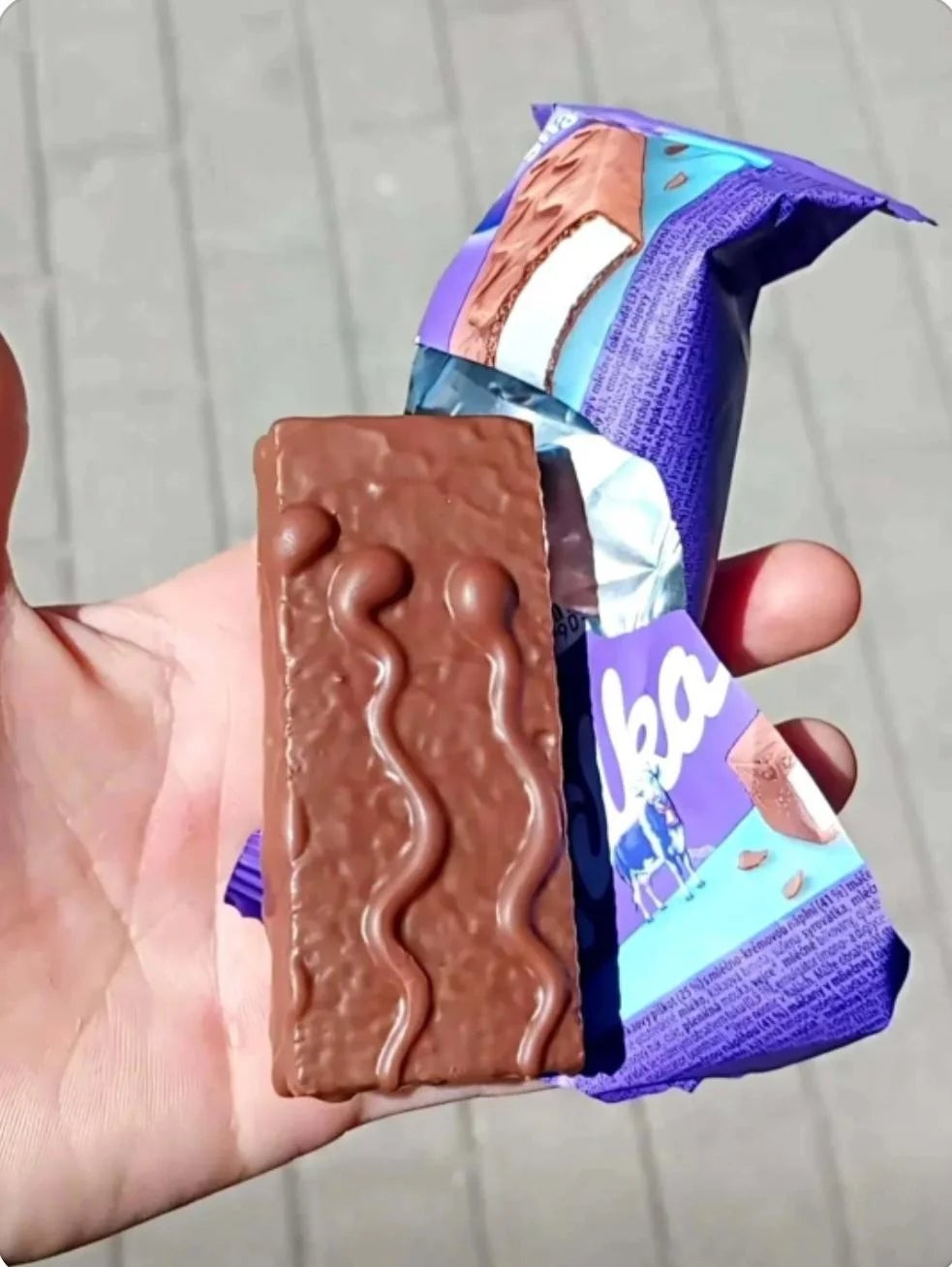 A manus  holds a Milka cocoa  barroom  with the wrapper partially open, showing a wavy cocoa  signifier   connected  the bar