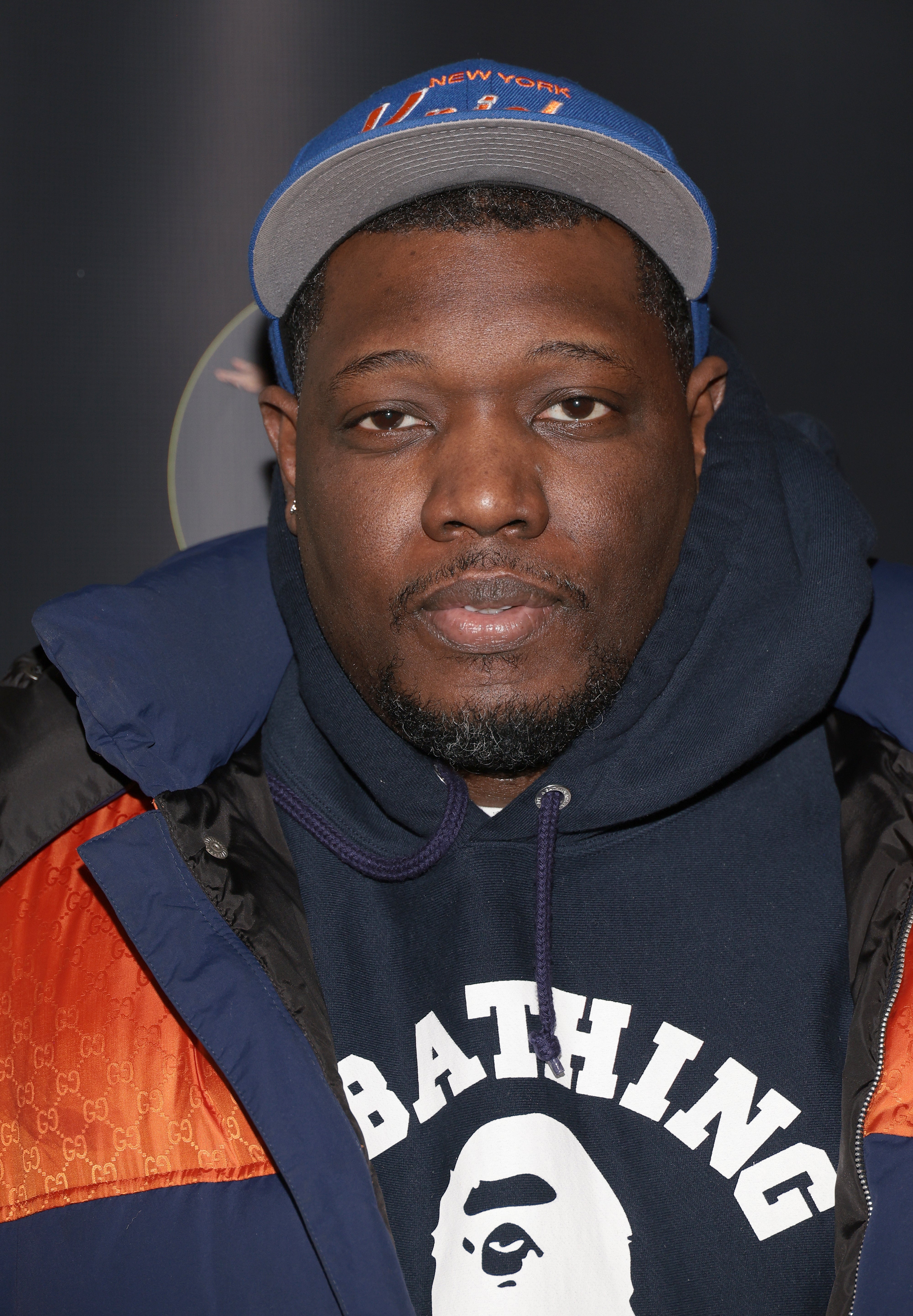 Michael Che wearing a puffy jacket, hoodie with a logo, and a baseball cap