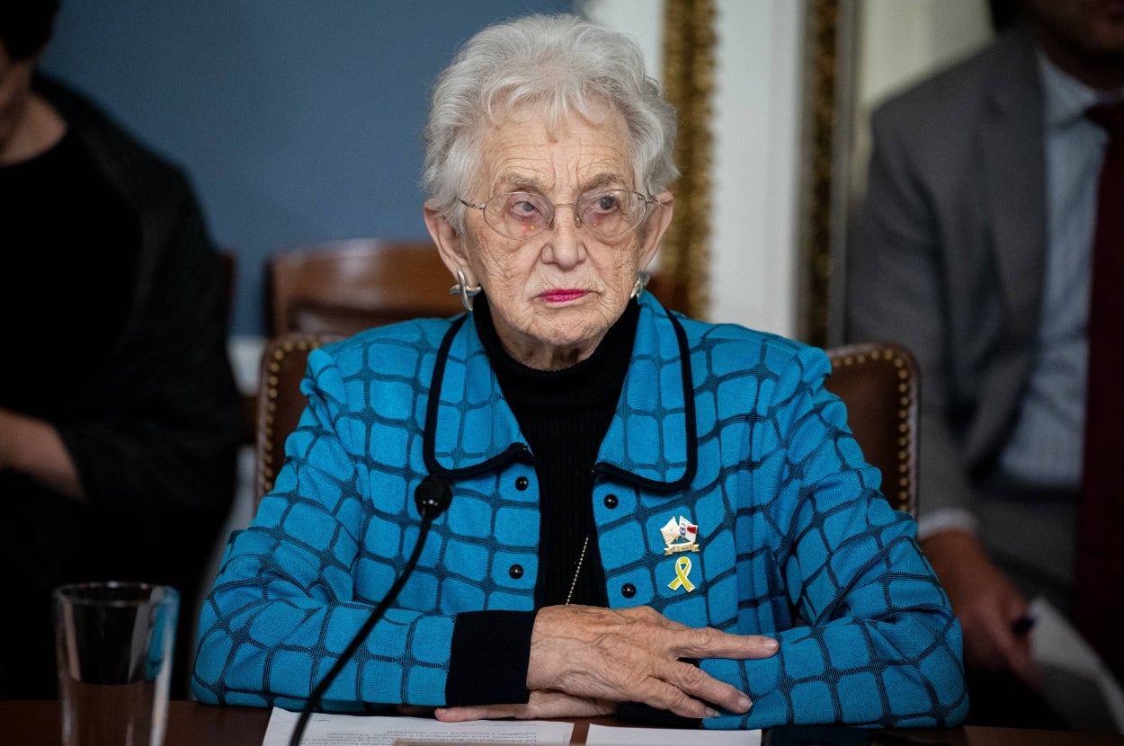 An elderly woman sits at a table, wearing a patterned jacket and pins. She looks attentively during a formal meeting