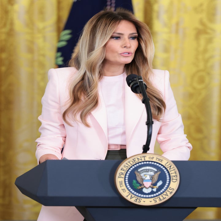A woman speaks at a podium with the U.S. presidential seal, wearing a tailored blazer and long wavy hair