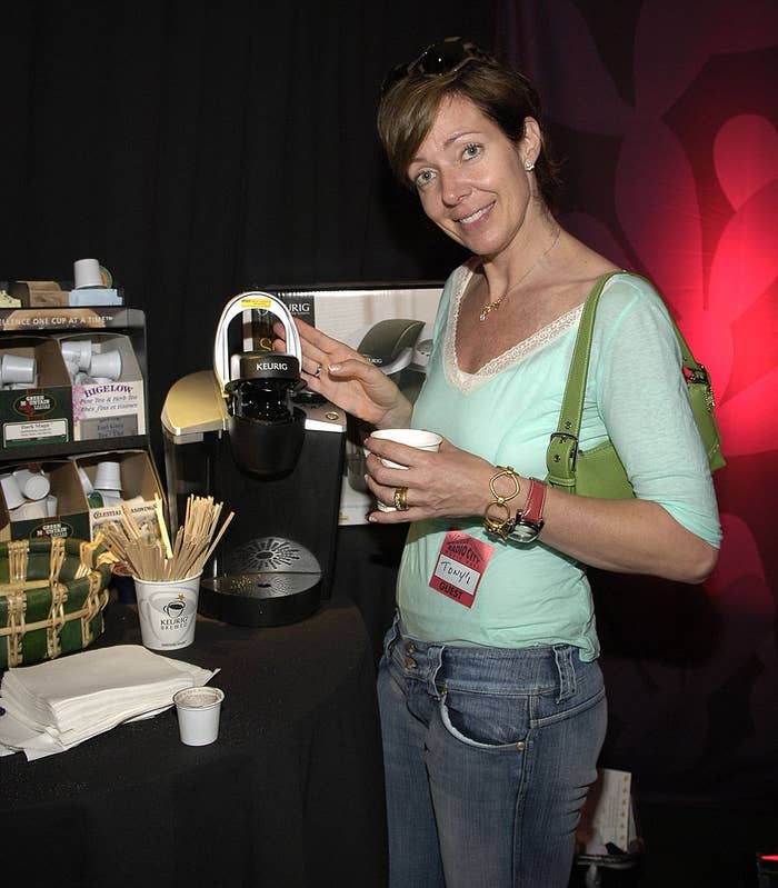 A idiosyncratic   stands by a java  instrumentality   display, holding a cup, wearing a casual greenish  apical  and jeans, with a greenish  purse implicit    the shoulder