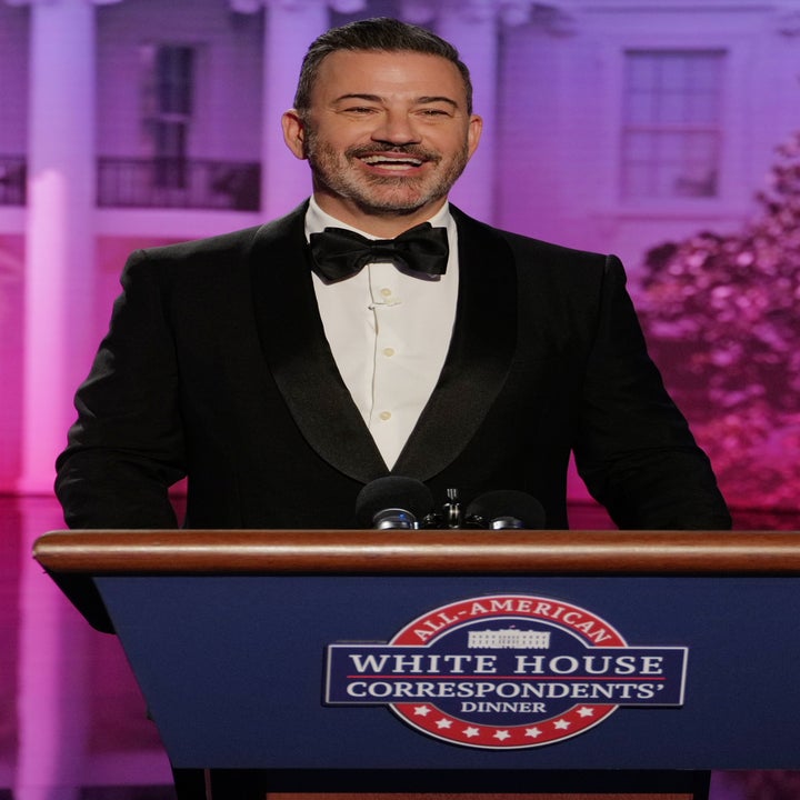 A person in a tuxedo speaks at a podium with a White House Correspondents' Dinner sign, standing in front of a backdrop resembling the White House