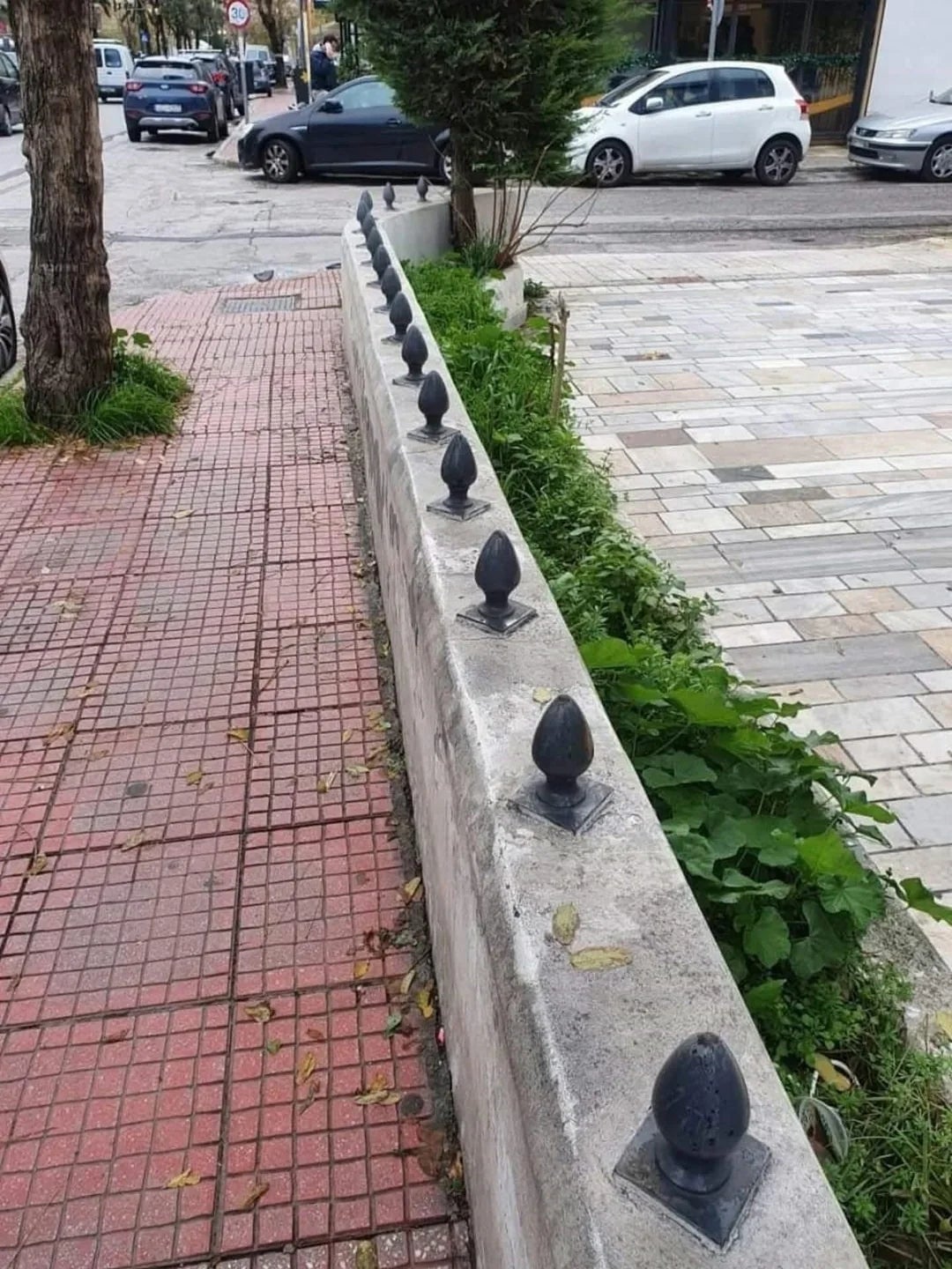 Low partition  topped with metallic  spikes lining a sidewalk; cars are parked connected  the street