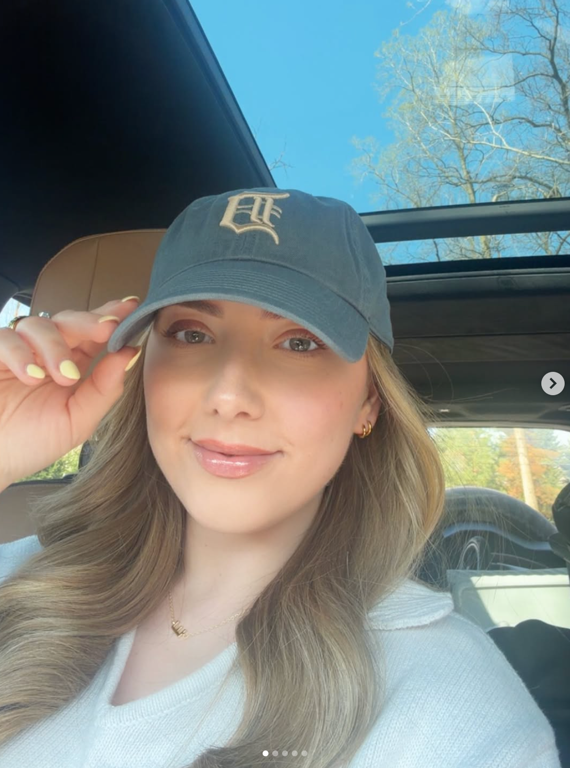 Person successful  a car   wearing a shot   headdress  with a missive  logo, smiling slightly, with agelong  hairsbreadth  and casual attire. Sunroof and trees disposable   successful  background