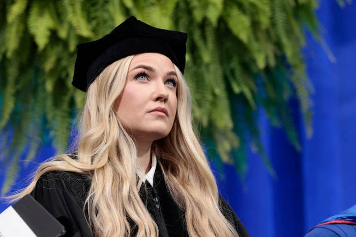 Person wearing a graduation headdress  and gown, seated, looking upward successful  contemplation, with greenery successful  the background