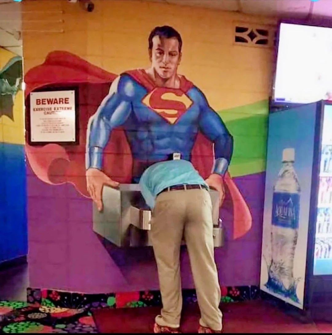 Person drinking from a h2o  fountain nether  a mural of a superhero with arms extended, creating a humorous optical illusion