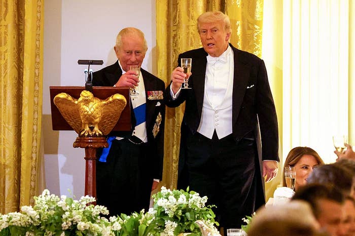 Two men in formal evening wear raise their glasses at a podium during an event. A woman is partially visible at the table below