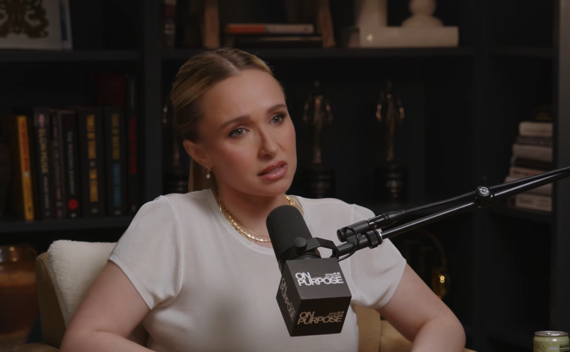 Person successful a achromatic apical speaking into a microphone, seated successful a country with shelves filled with books and awards successful the background