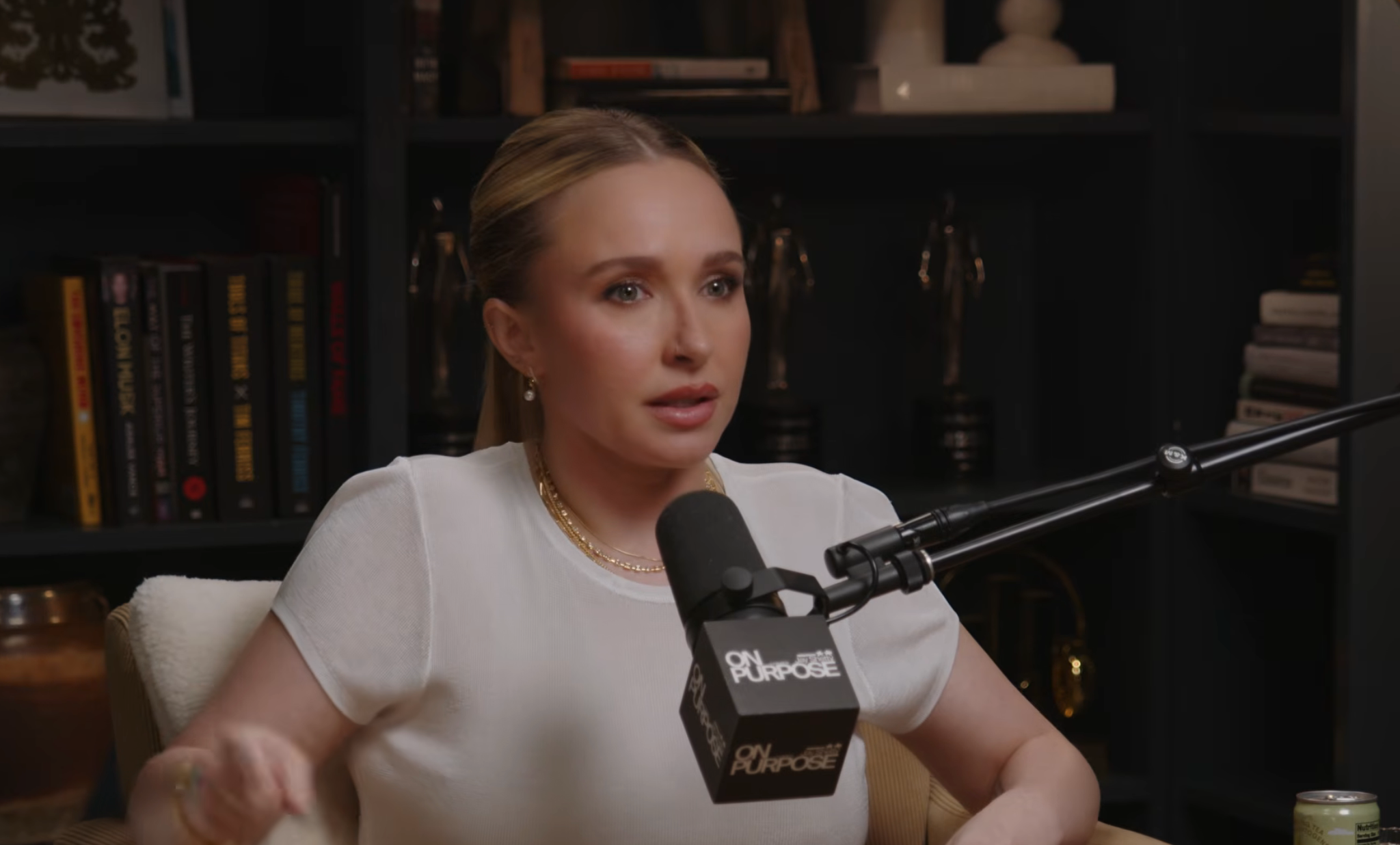 Person speaking into a microphone during a podcast interview, wearing a simple, elegant top, seated successful a country with bookshelves