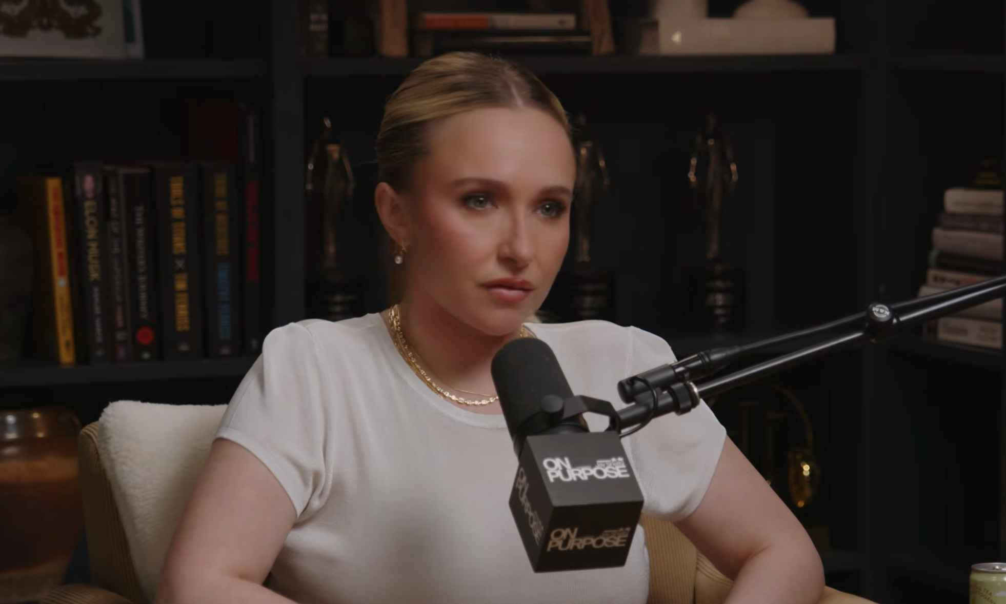 A idiosyncratic is sitting successful a podcast studio, wearing a achromatic apical and pearl necklace, speaking into a microphone. Books and awards are disposable successful the background