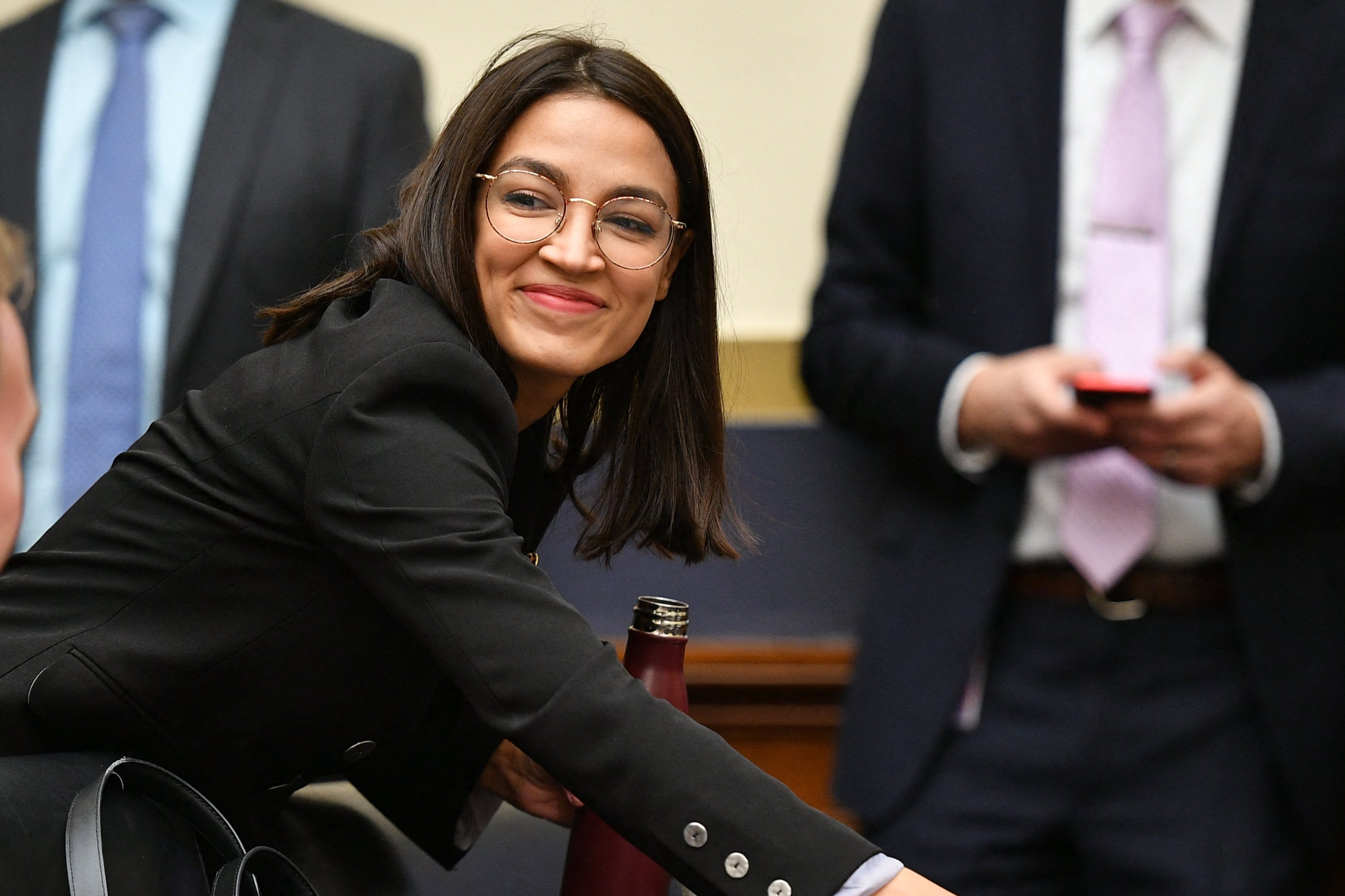 A idiosyncratic successful a suit and glasses smiles portion placing a cupful connected a array successful a ceremonial setting, surrounded by radical successful concern attire